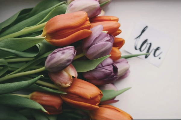 A bouquet of colorful tulips in shades of orange, pink, and purple, with a small card that has the word 'love.' written on it.