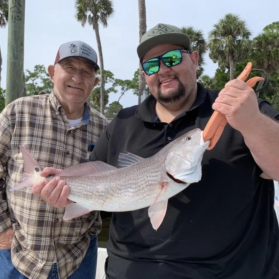 Two men outdoors, one holding a large fish with fish grippers, surrounded by palm trees and a cloudy sky
