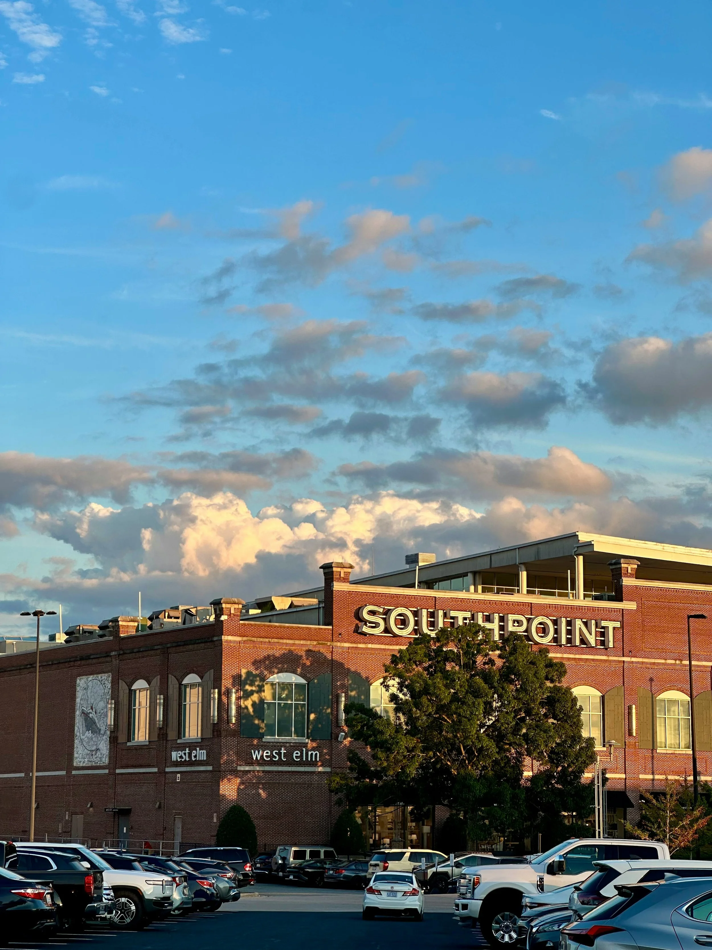 A brick building with a sign that says 'SOUTHPOINT' at the top, a parking lot with several cars in front, and a blue sky with clouds overhead.