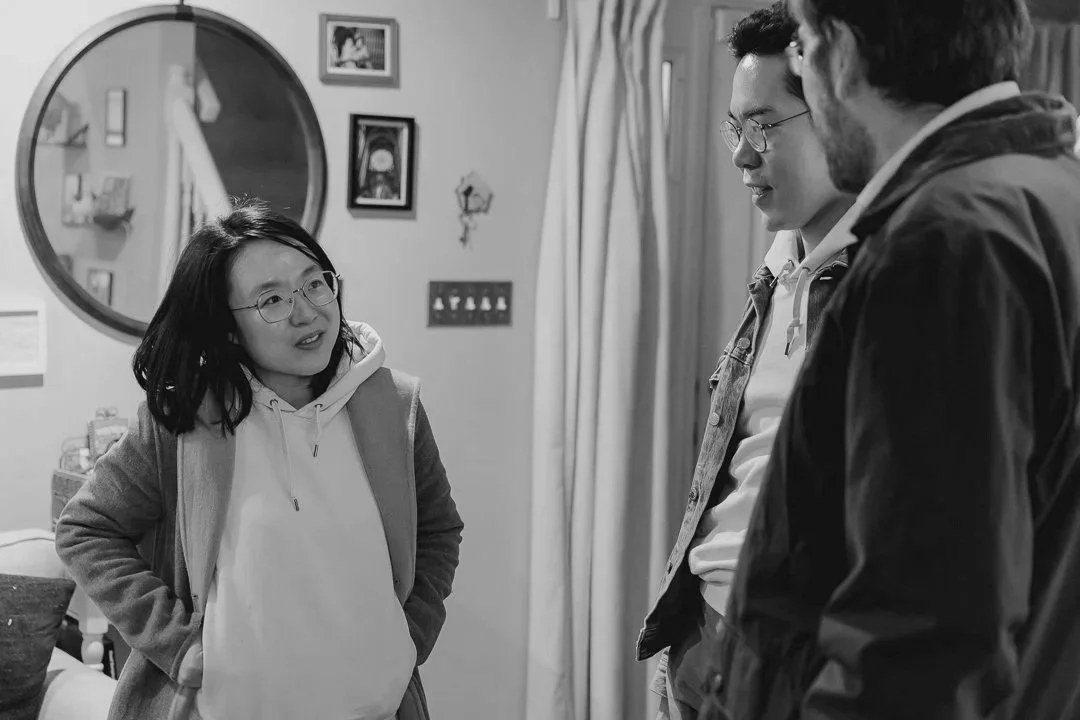 3 people in a living room having a conversation. A woman in a hoodie, and a coat is facing two adults, one of whom is wearing glasses and the other has a beard and glasses. The room has a mirror, framed photos, and curtains in the background.