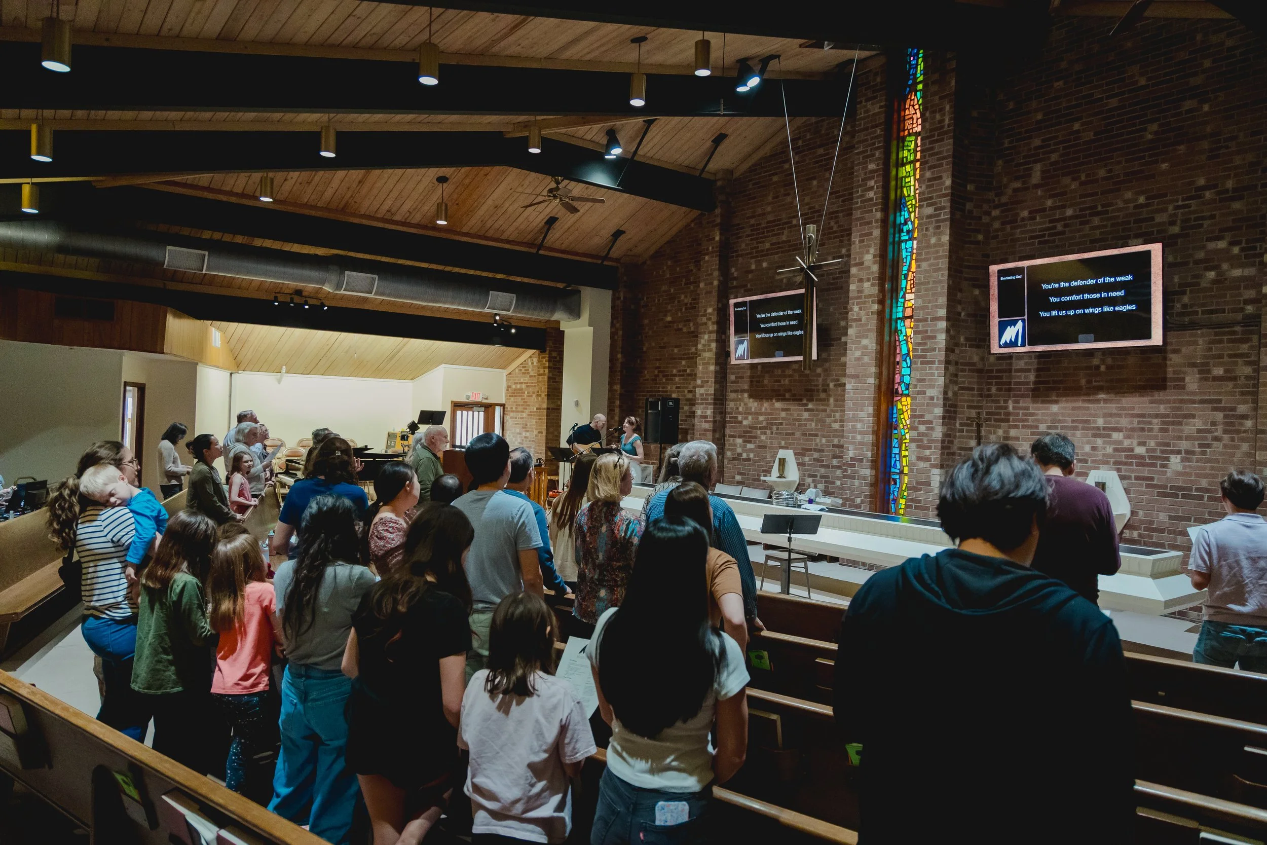 A congregation gathered in a church during a service or event, standing in front of the altar with some people singing or praying, while others listen. The interior features a high wooden ceiling, brick walls, and two screens displaying song lyrics.
