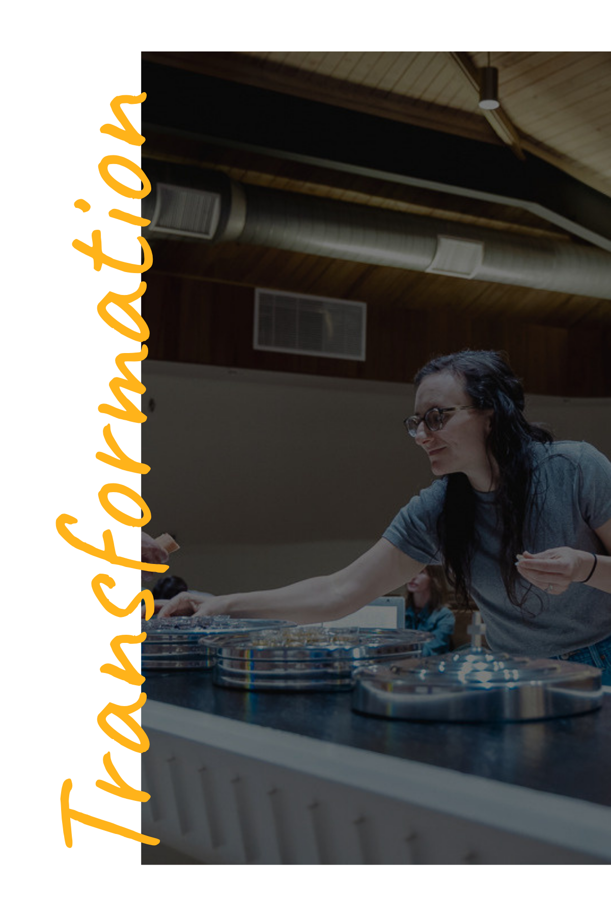 A woman with long dark hair and glasses reaching across a table with game pieces or board game tokens in a room with wooden ceilings and visible ductwork.