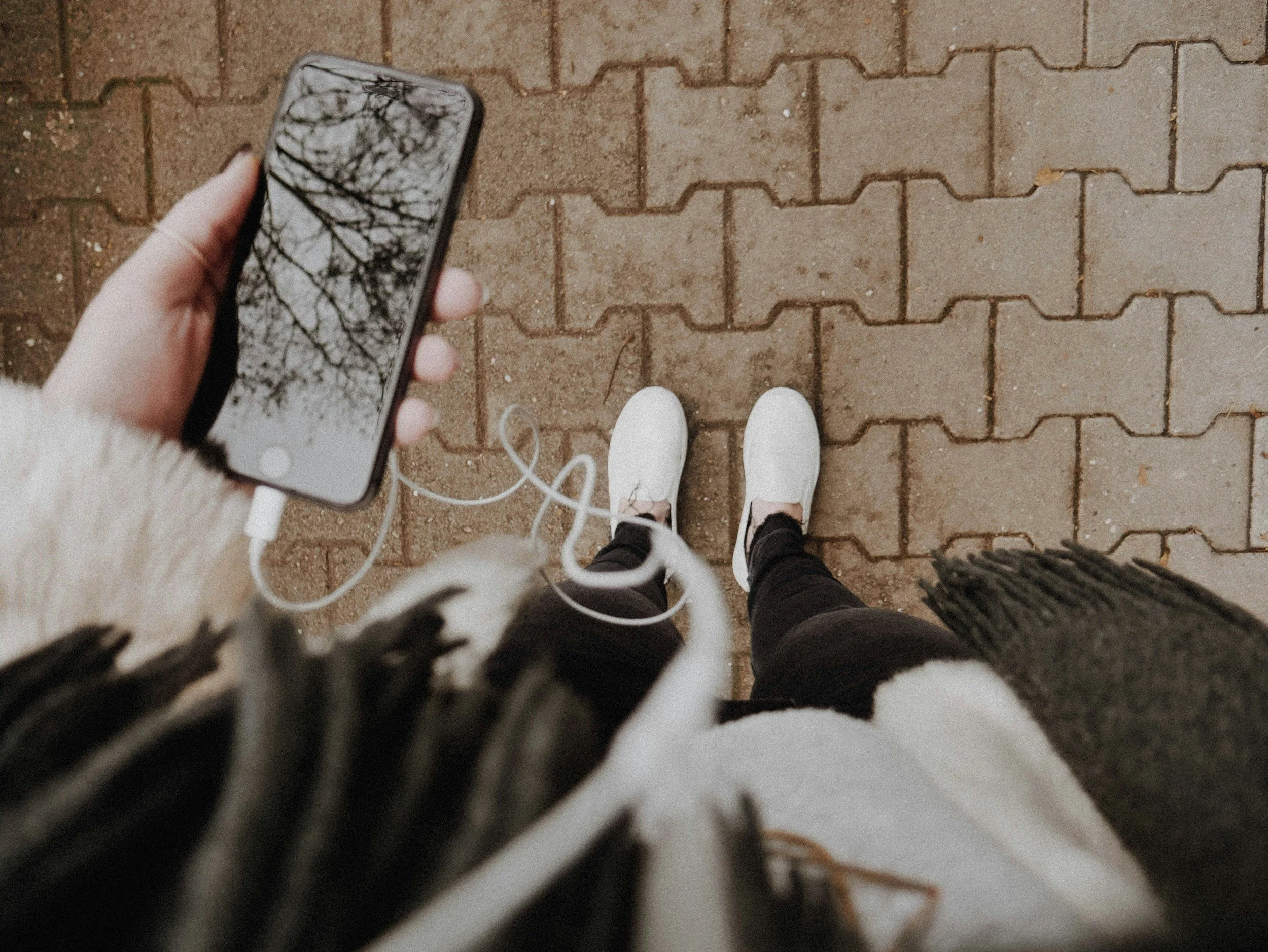 A person's feet in white sneakers standing on a brick sidewalk, holding a smartphone showing a reflection of tree branches.