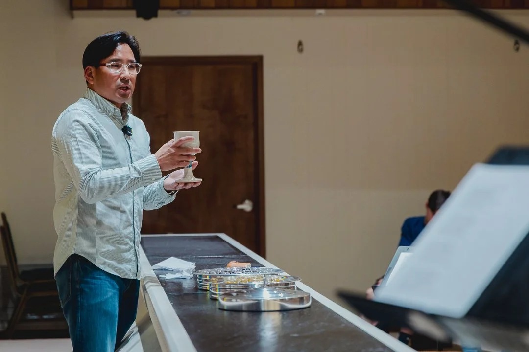 A man with black hair, glasses, wearing a light-colored striped shirt, standing at a counter, holding a chalice and speaking to an audience.
