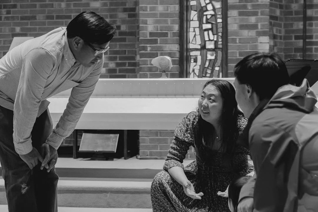 Three people engaged in a conversation inside a church or a similar building. One woman is sitting on the floor, speaking to two men who are leaning toward her. The setting includes brick walls and stained glass windows.