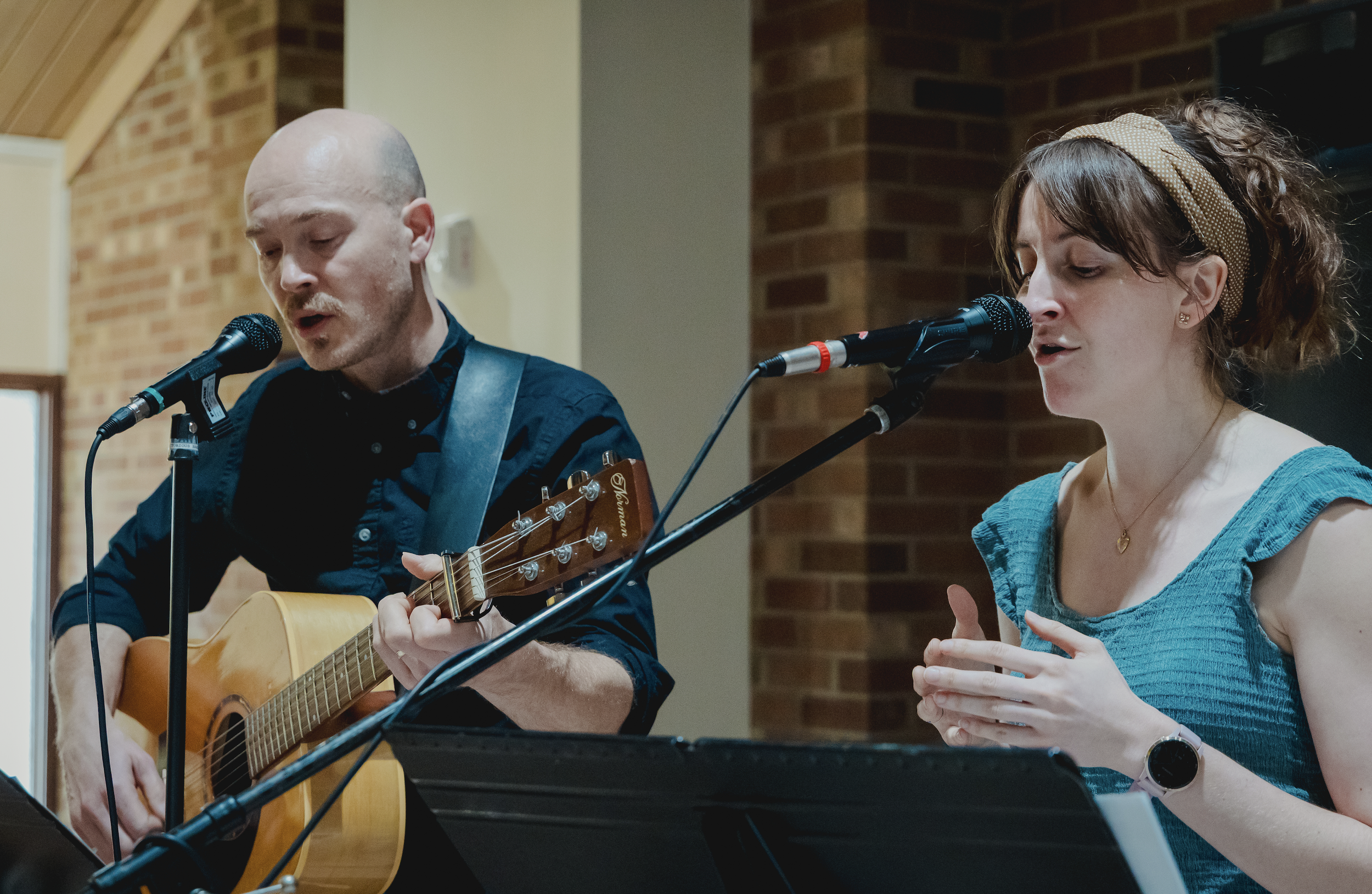 A man and a woman singing into microphones while the man is playing an acoustic guitar in a room with brick walls.