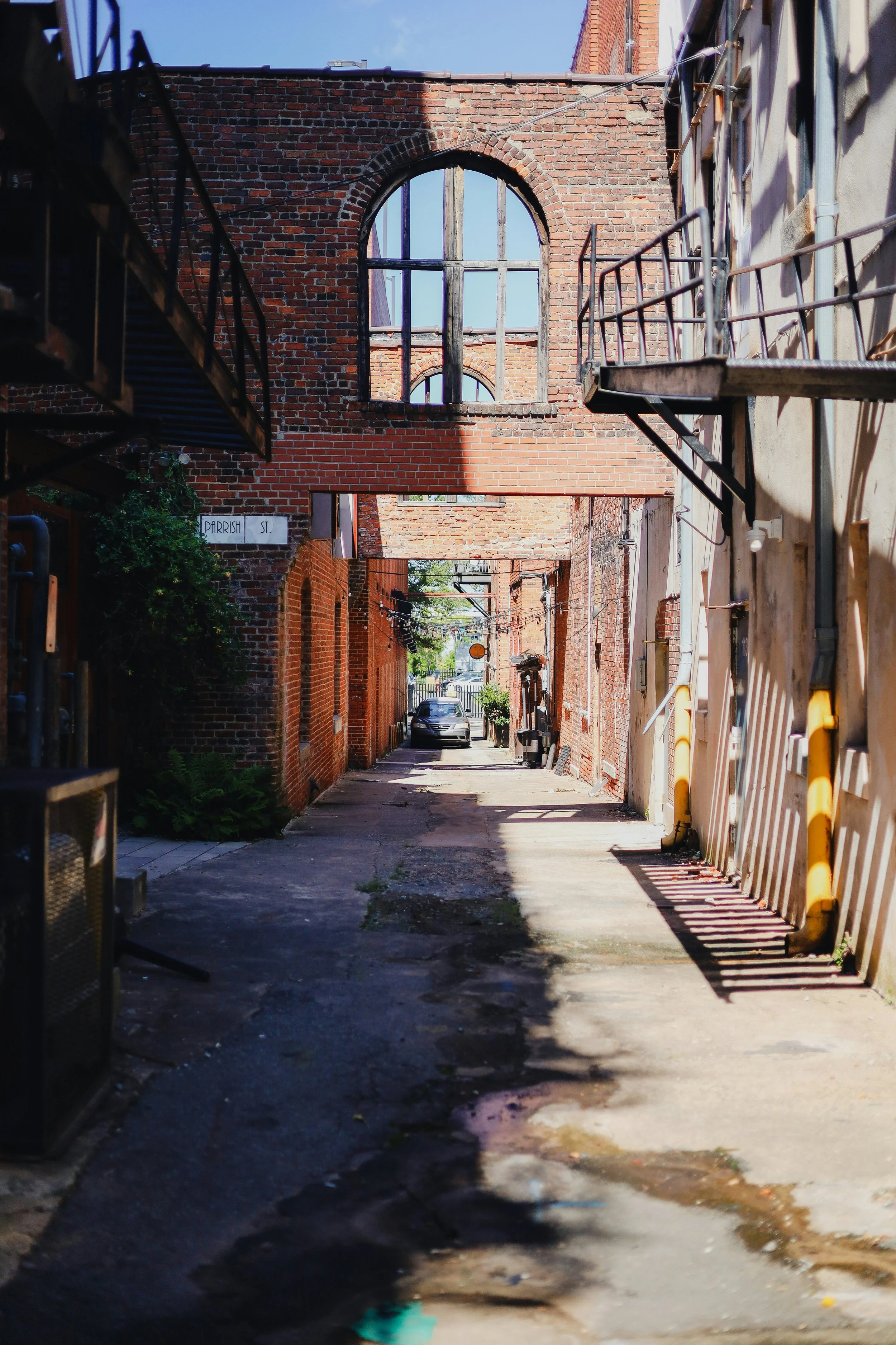 A narrow alleyway in durham between brick and concrete buildings, with a car parked in the distance, fire escapes on both sides, and an arched window in a brick wall overhead.