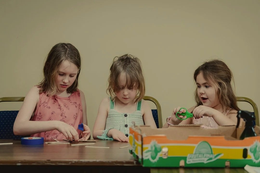 Three young girls sitting at a table playing with wooden sticks and toy scissors.