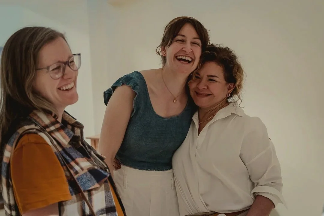 Three women laughing together indoors, one woman in a plaid shirt and glasses, another in a blue top, and the third in a white blouse.