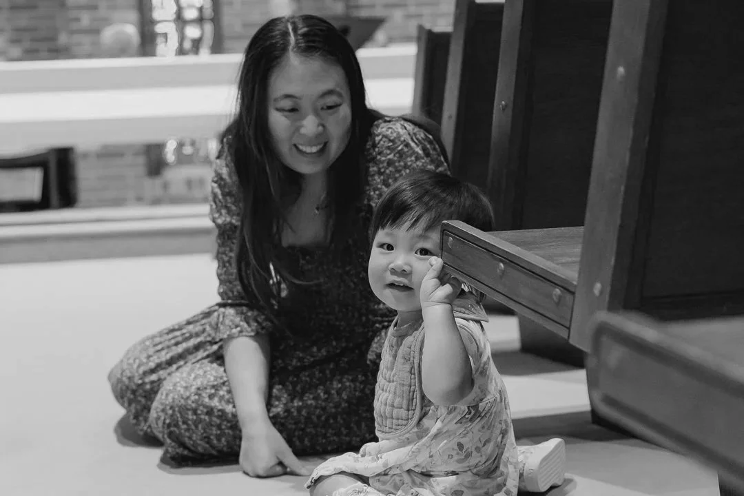 A woman and a young child sitting on the floor, with the woman smiling and the child holding a wooden block near their face.