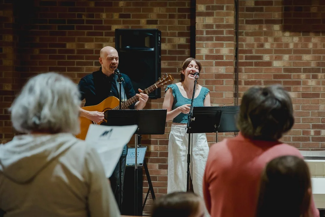 Two musicians, a man playing guitar and singing and a woman singing, perform for an audience in a room with brick walls.