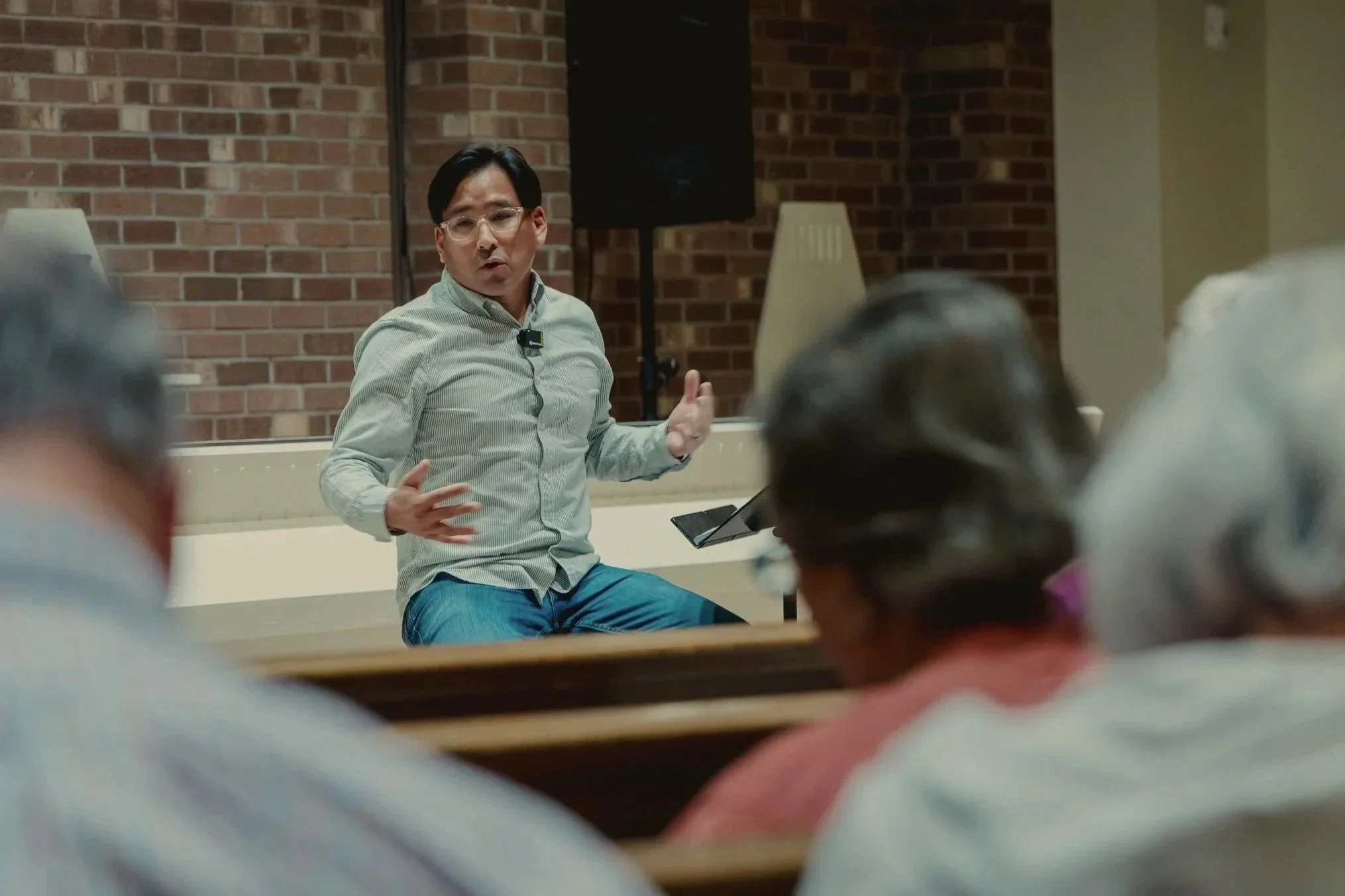 Didi preaching at our sunday afternoon service at missio. Man wearing glasses, speaking in a meeting with a brick wall in the background, seated at a table with people around him.