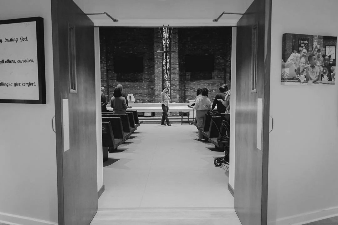 View through open doors into a church or chapel with people standing and sitting, some praying or reading, and a large cross in the background.