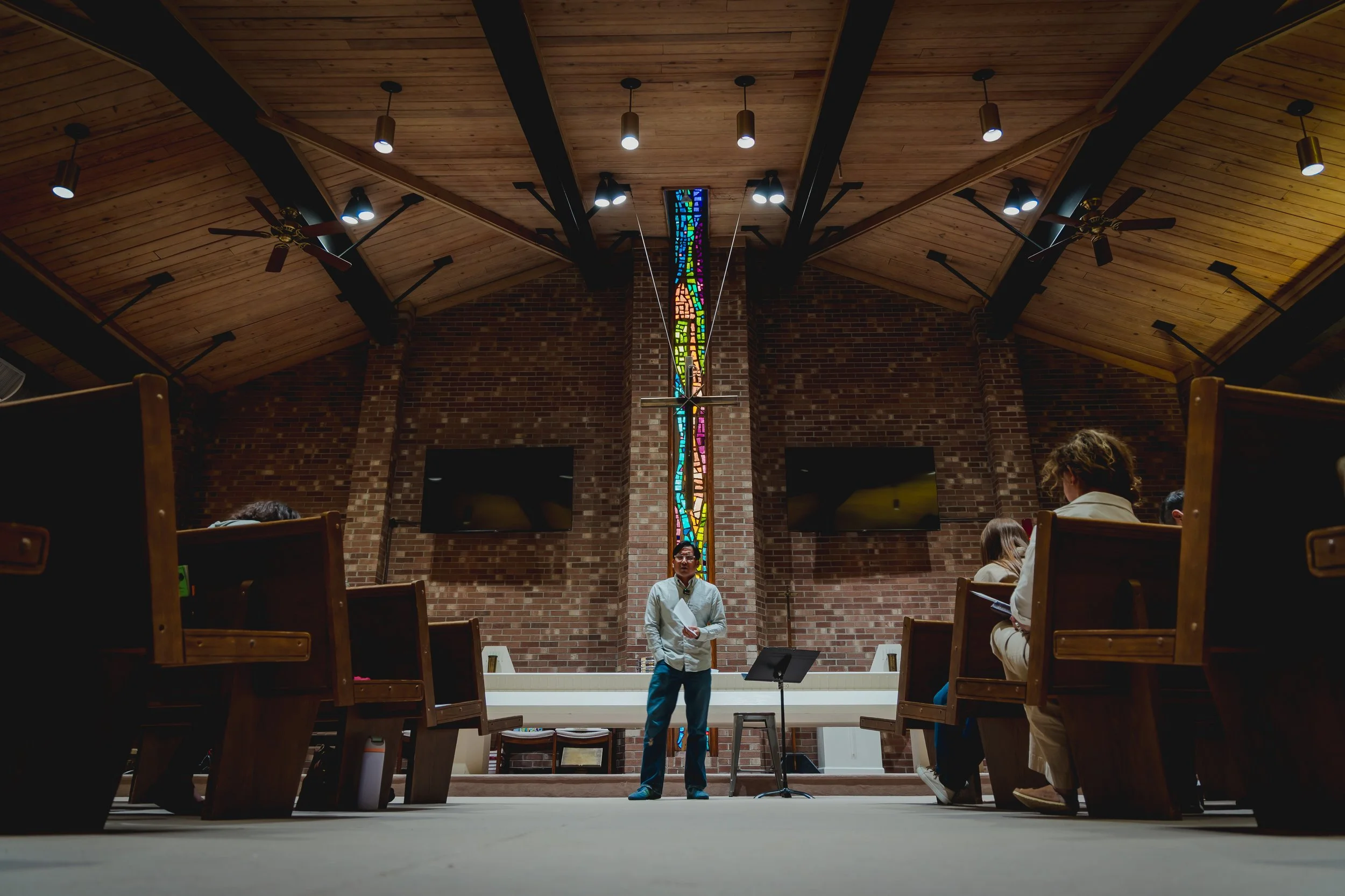 Interior of a church or auditorium with a wooden ceiling, brick walls, and stained glass window. Several people are seated and standing, with one man speaking at a podium.