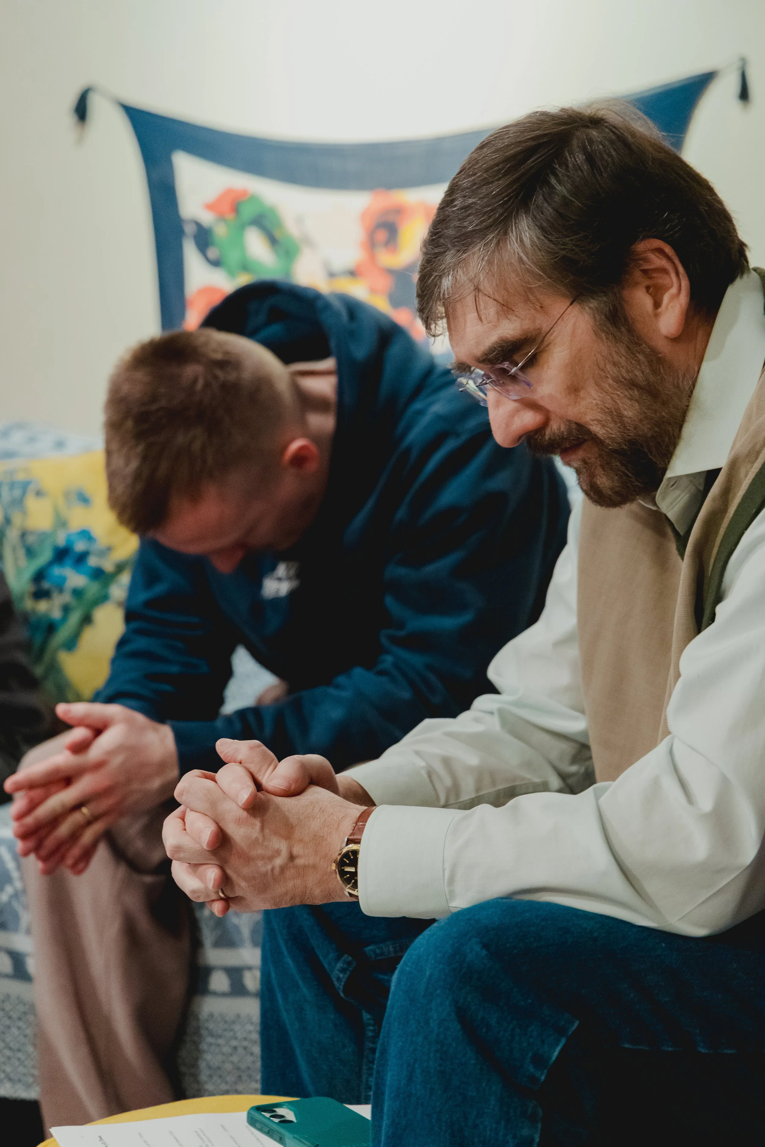 A group of people at missio group in durham sitting in a row with their hands clasped, involved in prayer. One man with glasses and a beard is most prominently featured, with other individuals in the background.