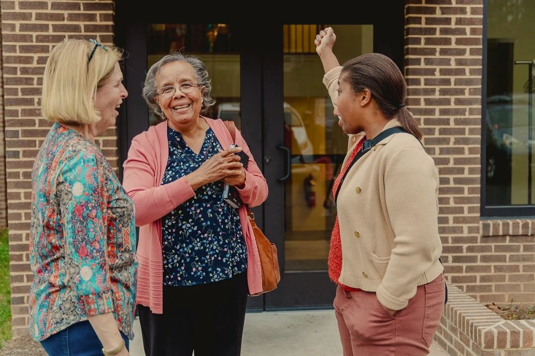Three women standing outside after Missio durham's sunday service and talking, one woman raising her arm in excitement.