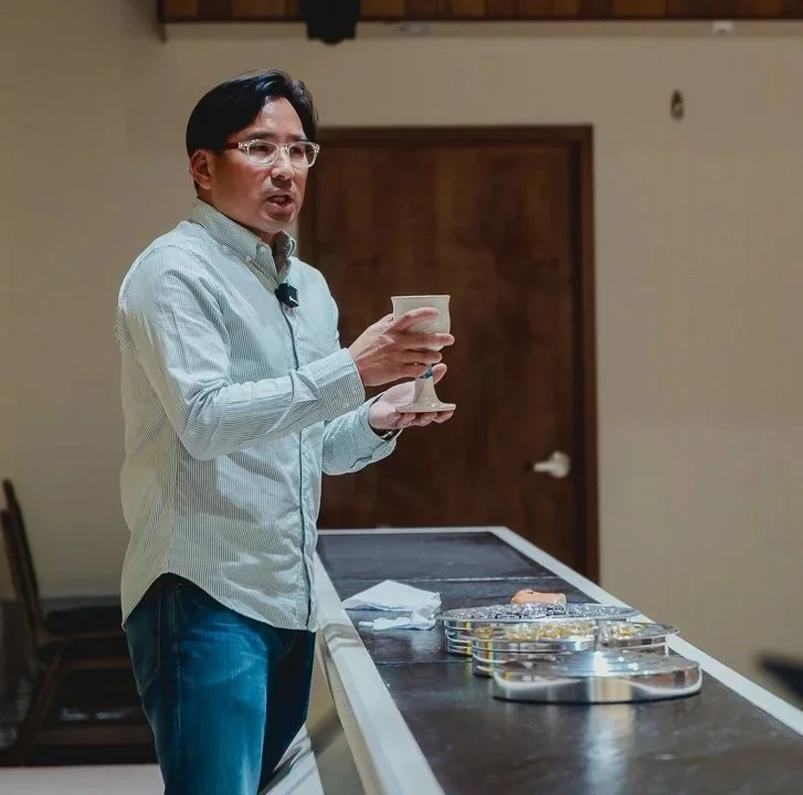 Man speaking and holding a cup and saucer in a room with a table and sealed snacks.