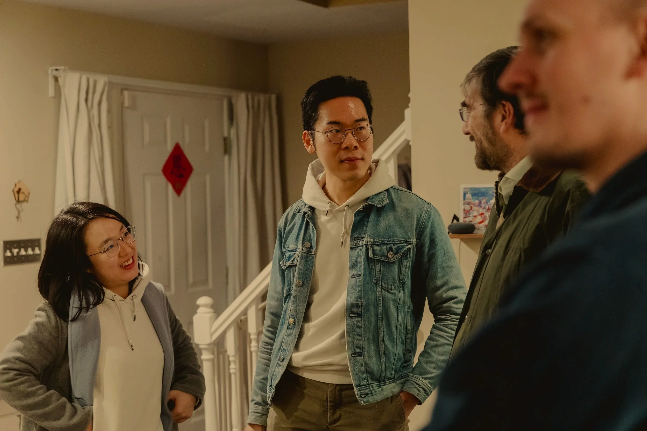 A group of four diverse people engaging in conversation inside a home, with a staircase and holiday decorations in the background.