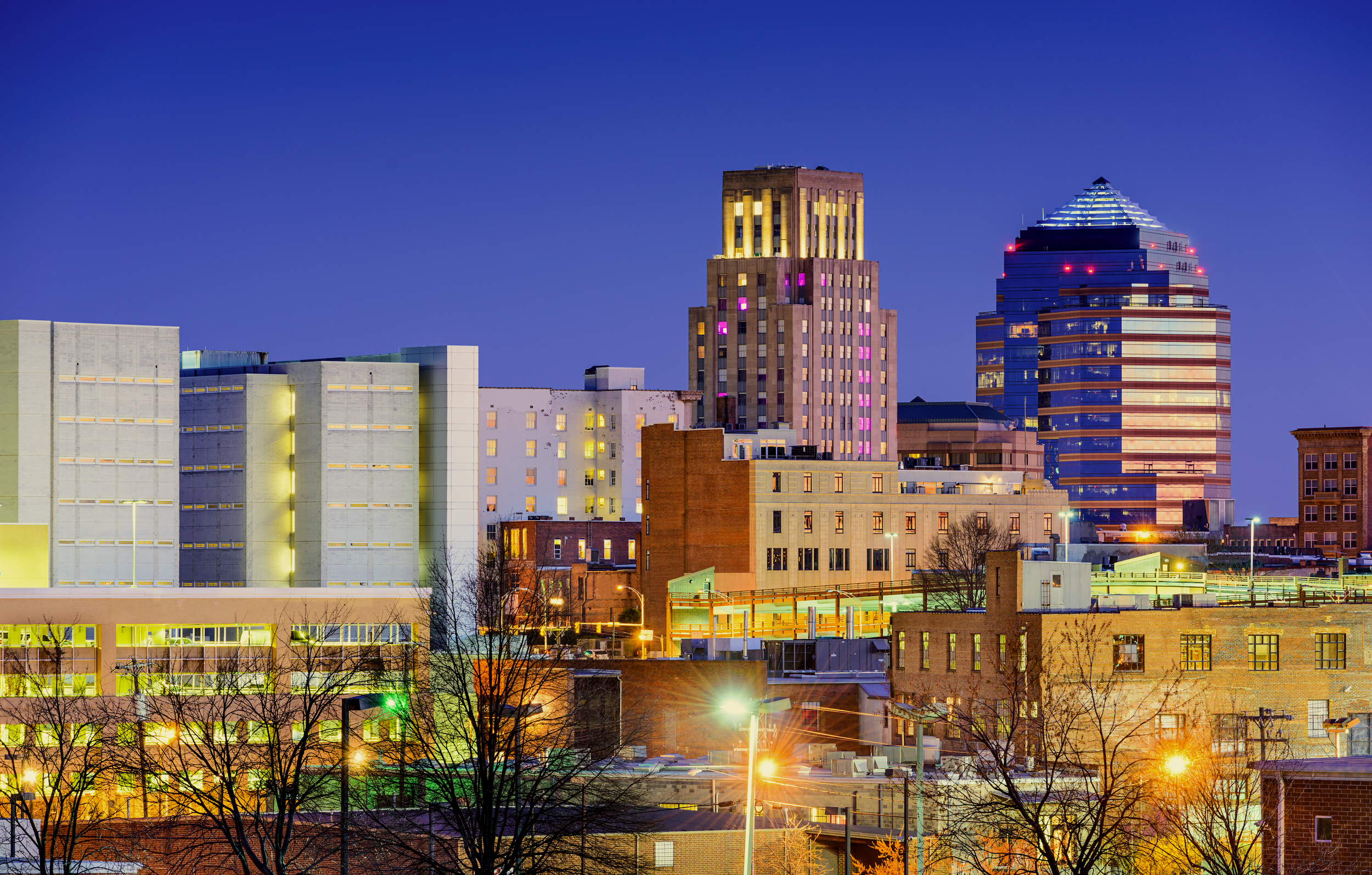 Nighttime city skyline of Durham, NC, with illuminated buildings against a clear blue sky, including a tall Art Deco-style skyscraper on the left and a modern glass tower on the right, with streetlights and bare tree silhouettes in the foreground.
