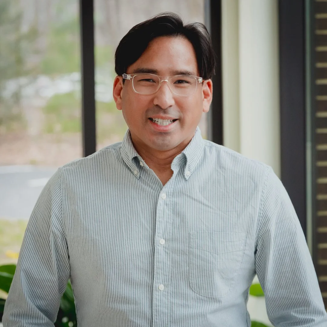 A smiling man with black hair and glasses, wearing a light striped button-up shirt, standing indoors near large windows with a blurred outdoor background.