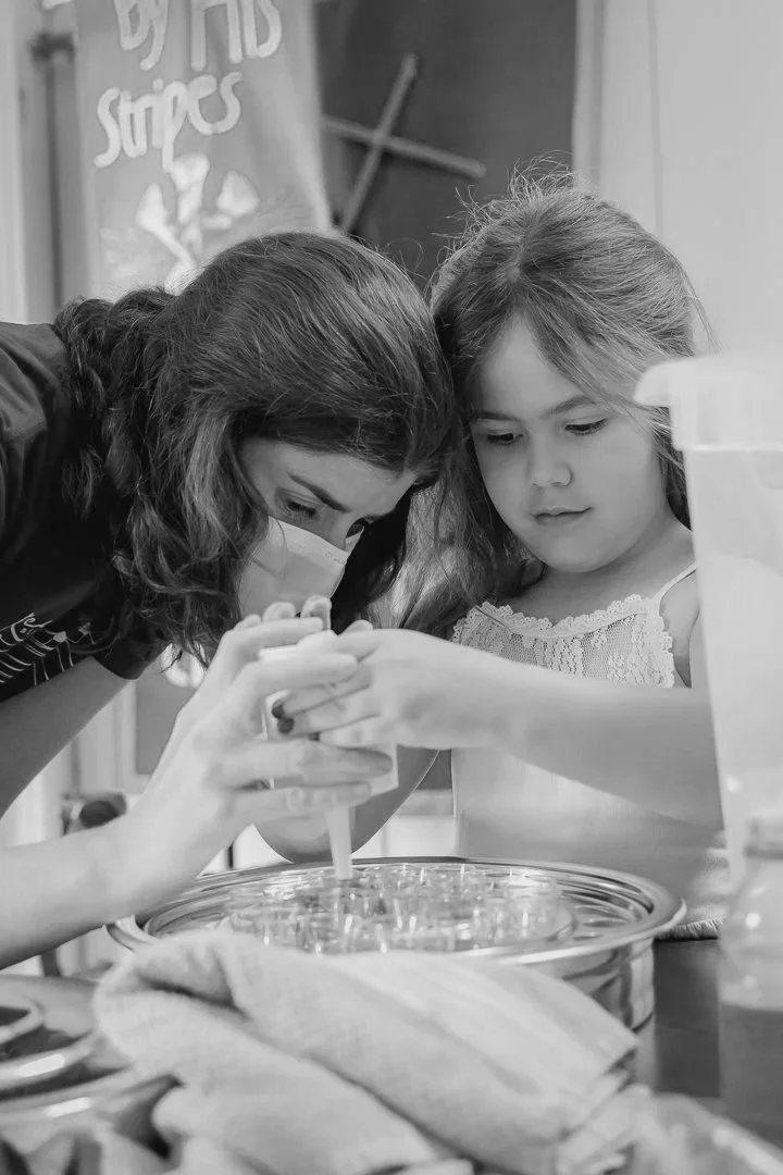 Two young girls, one with a face mask, are setting up communion. They are leaning over a bowl or container, with one girl pouring a liquid or substance from a bottle.