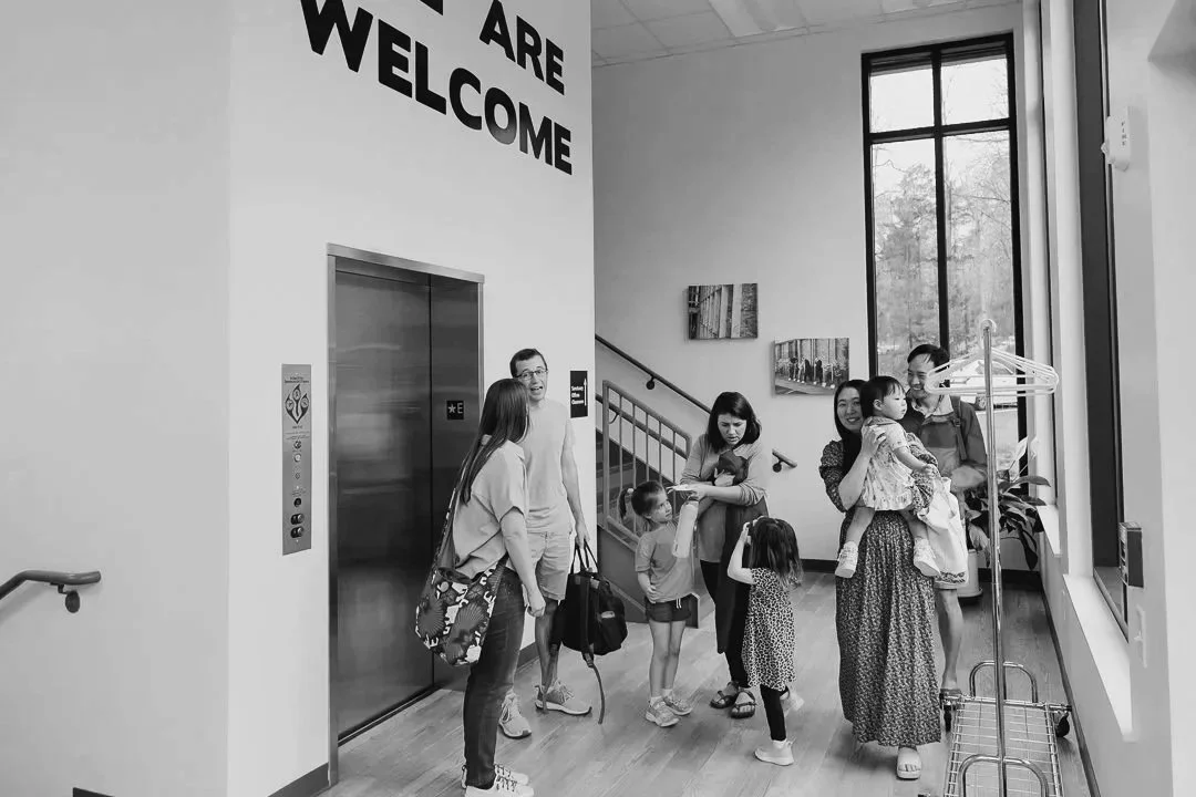 Group of men, women and children waiting in a modern building lobby near an elevator, with some talking and others looking at their phones, next to a large window with a coat rack nearby.