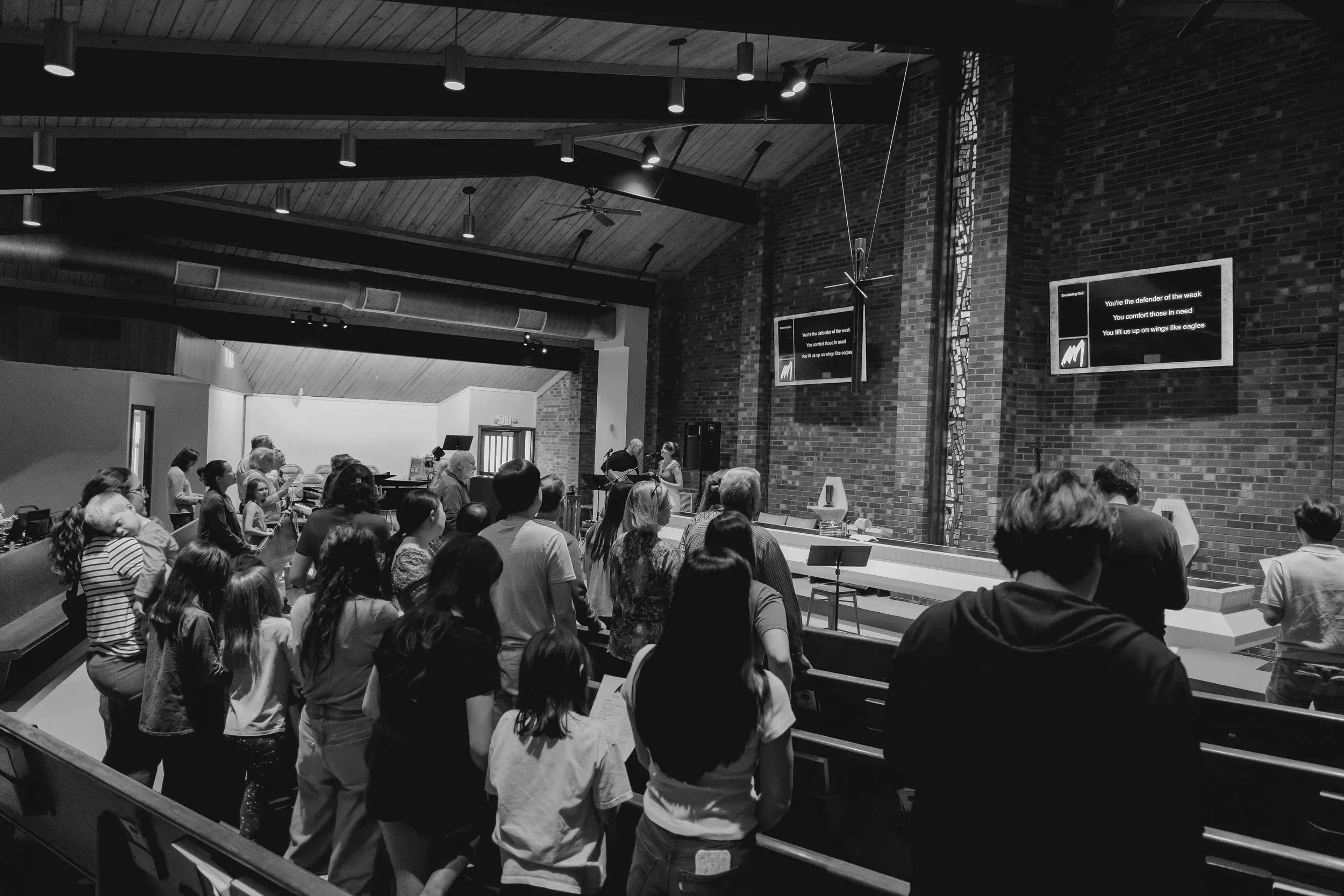 People gathered for Missio's sunday service in Durham, with some standing and others seated. The room has a high, wooden ceiling with hanging light fixtures, brick walls, and digital screens displaying song lyrics.