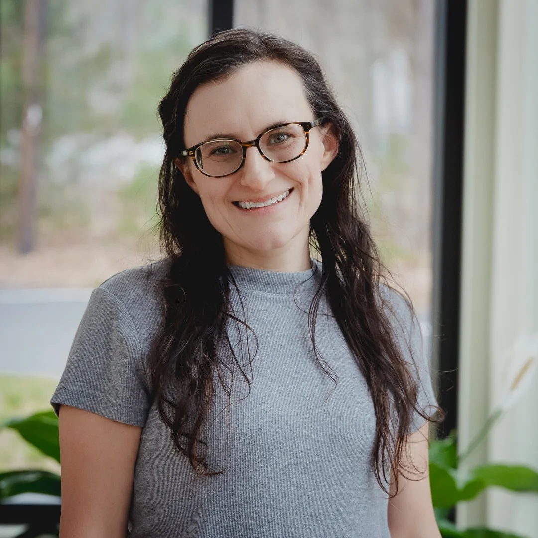 A woman with long dark hair, glasses, and a gray t-shirt smiling indoors near a window with green trees outside.