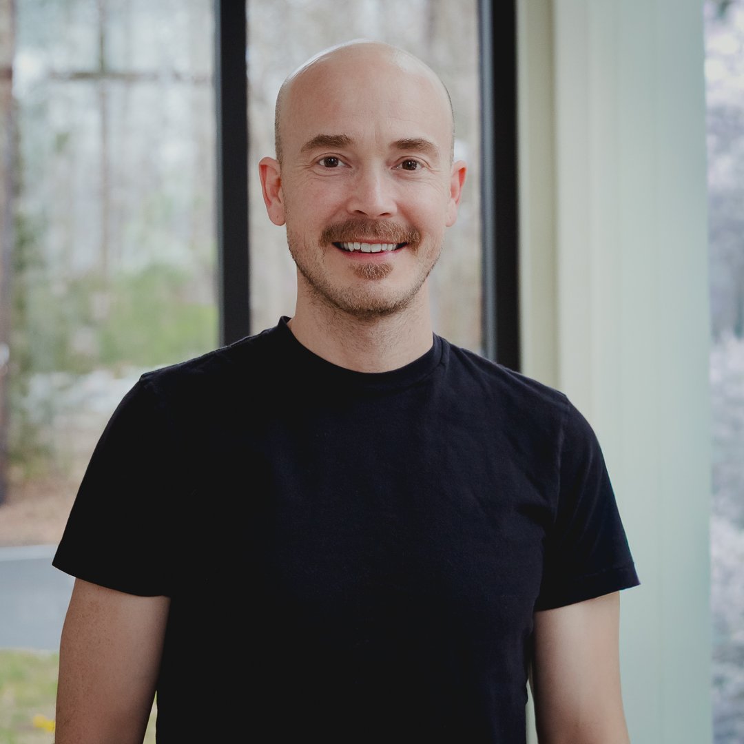 A smiling man with a bald head and a short beard, wearing a black t-shirt, standing indoors near large windows during the daytime.