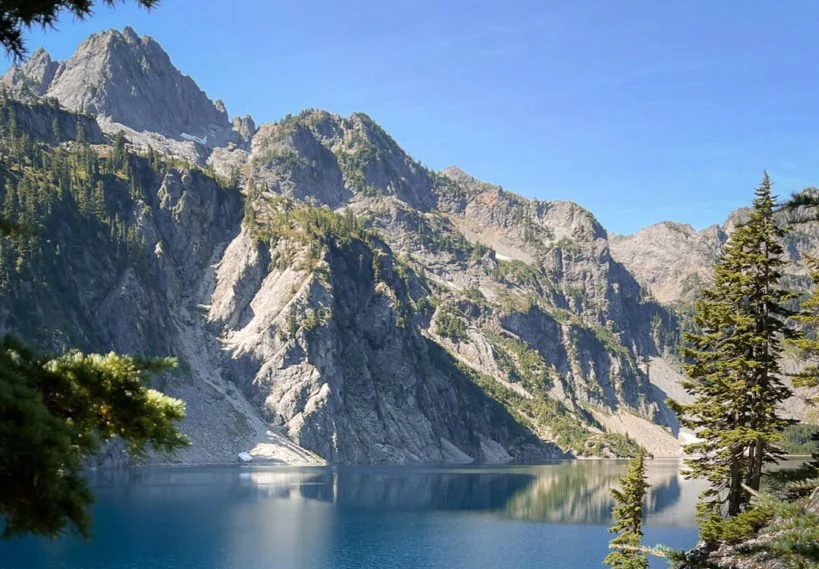 Scenic view of a lake surrounded by rugged mountains with sparse vegetation and a few tall evergreen trees in the foreground, under a clear blue sky.
