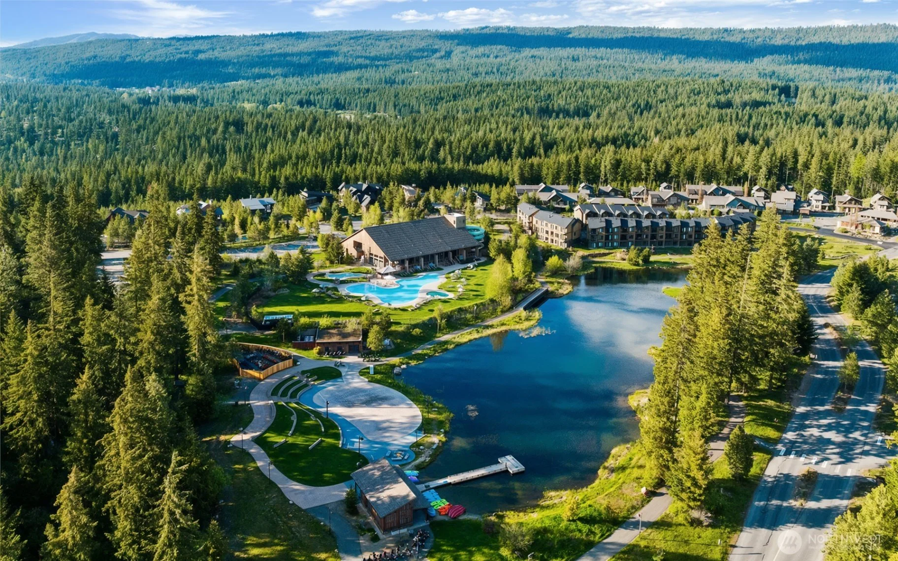 Aerial view of a resort surrounded by dense forest, with a large building featuring a swimming pool, a pond, walking paths, and multiple smaller buildings with cabins and parking areas.