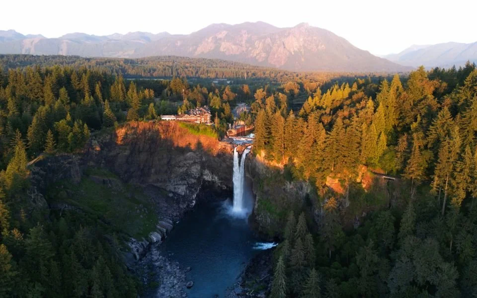 Aerial view of a tall waterfall flowing into a river, surrounded by dense green forest with distant mountains in the background, during sunset.