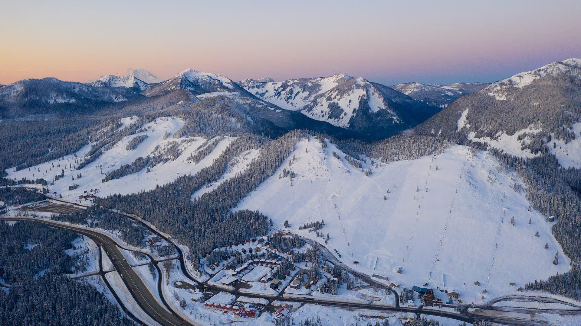 A snowy mountain landscape at dusk with snow-covered peaks, forested slopes, and a ski resort on the lower hills, including ski slopes and buildings. A winding highway runs through the bottom left corner.
