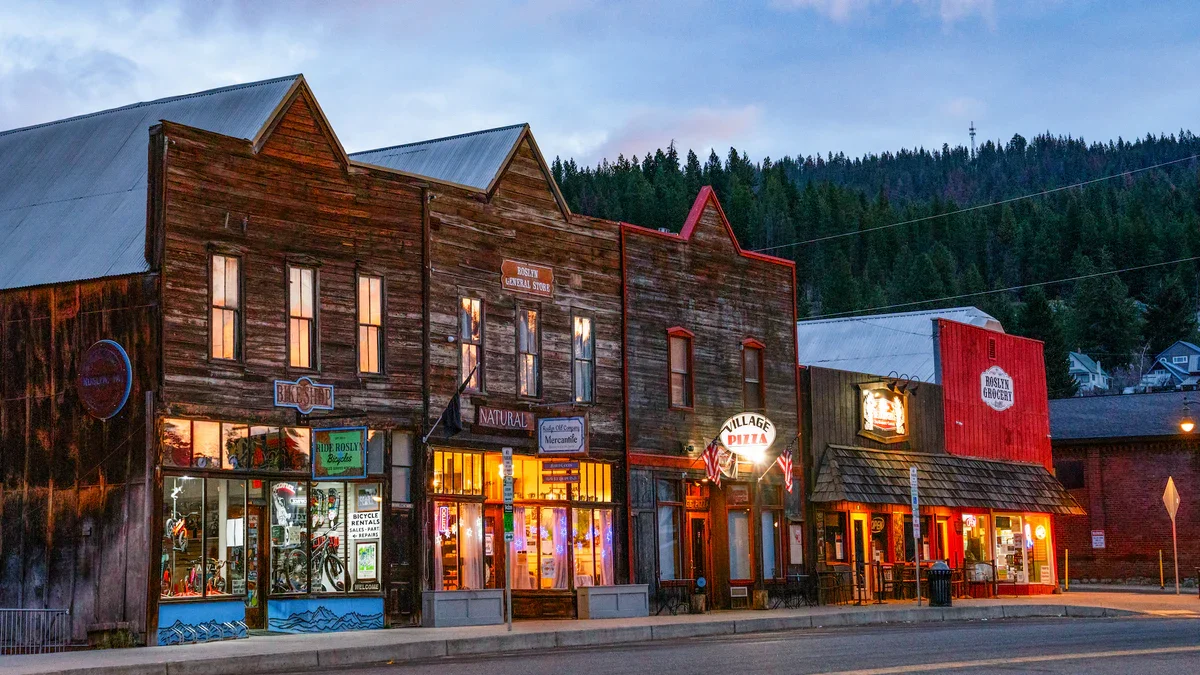 A row of rustic wooden storefronts in a small town, with a bike shop, grocery store, and pizza restaurant, under a cloudy sky with forested hills in the background.