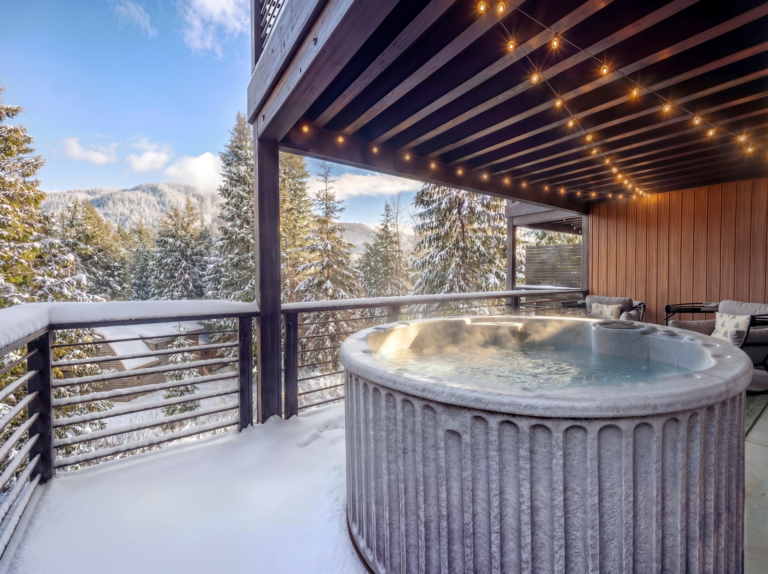 An outdoor balcony with a hot tub, surrounded by snow and overlooking a snowy forest with evergreen trees and mountain hills under a clear blue sky with some clouds. The balcony has string lights hanging from the ceiling and wood-paneled walls.