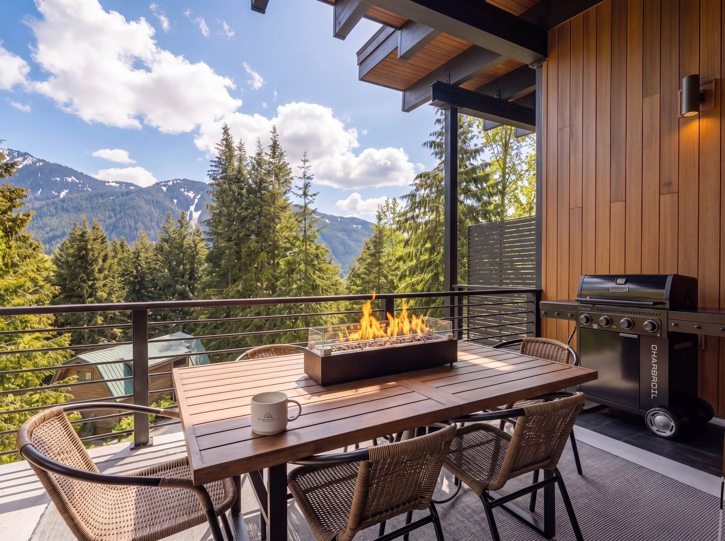 FIFA World Cup 2026 Seattle Airbnb Balcony with a wooden table, four chairs, a gas grill, and a modern fire pit, overlooking a forest and mountains under a partly cloudy sky.