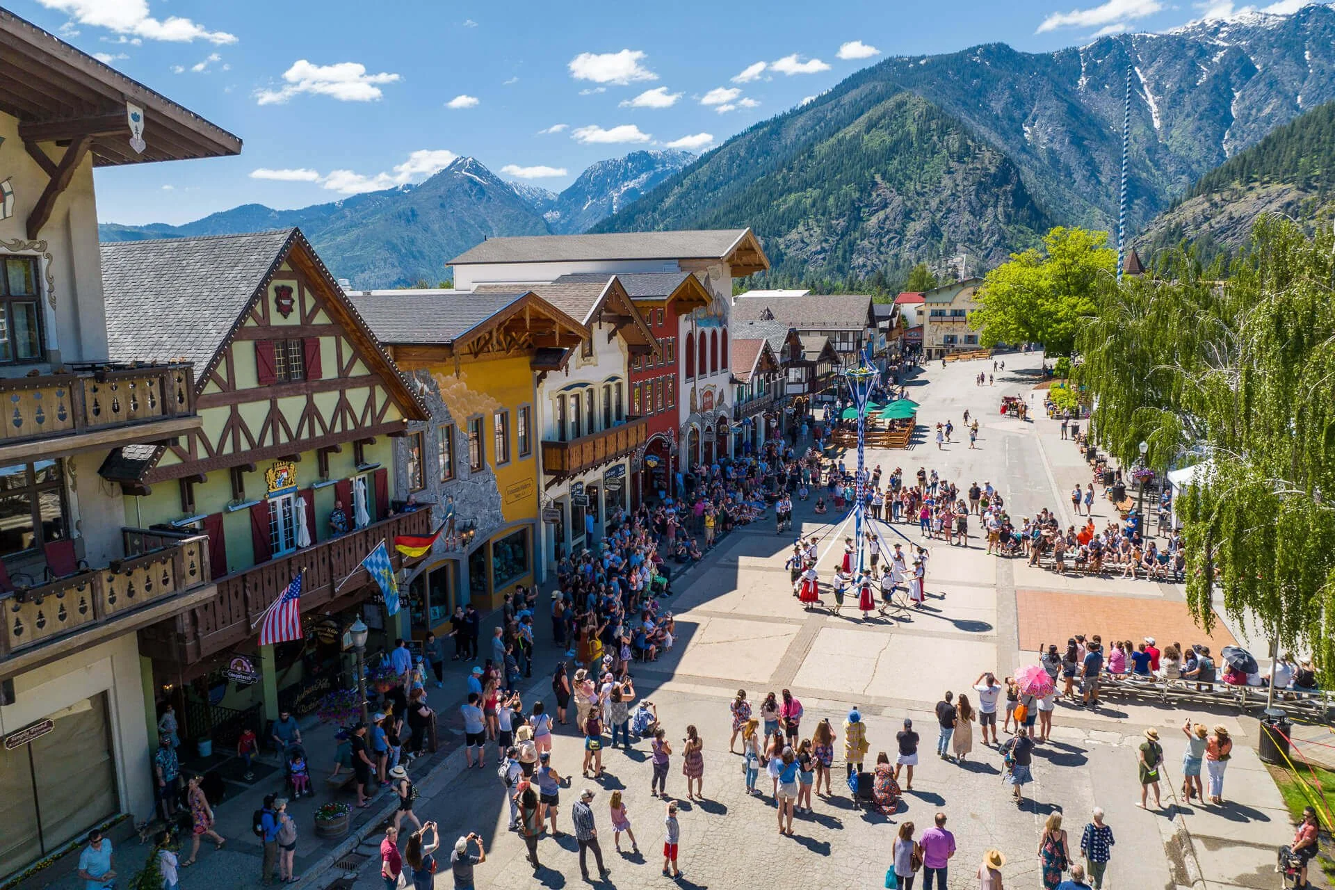 A busy outdoor scene in a mountain town square with colorful buildings, a crowd of people, and a flagpole with a blue and white striped flag. Snow-capped mountains are in the background.