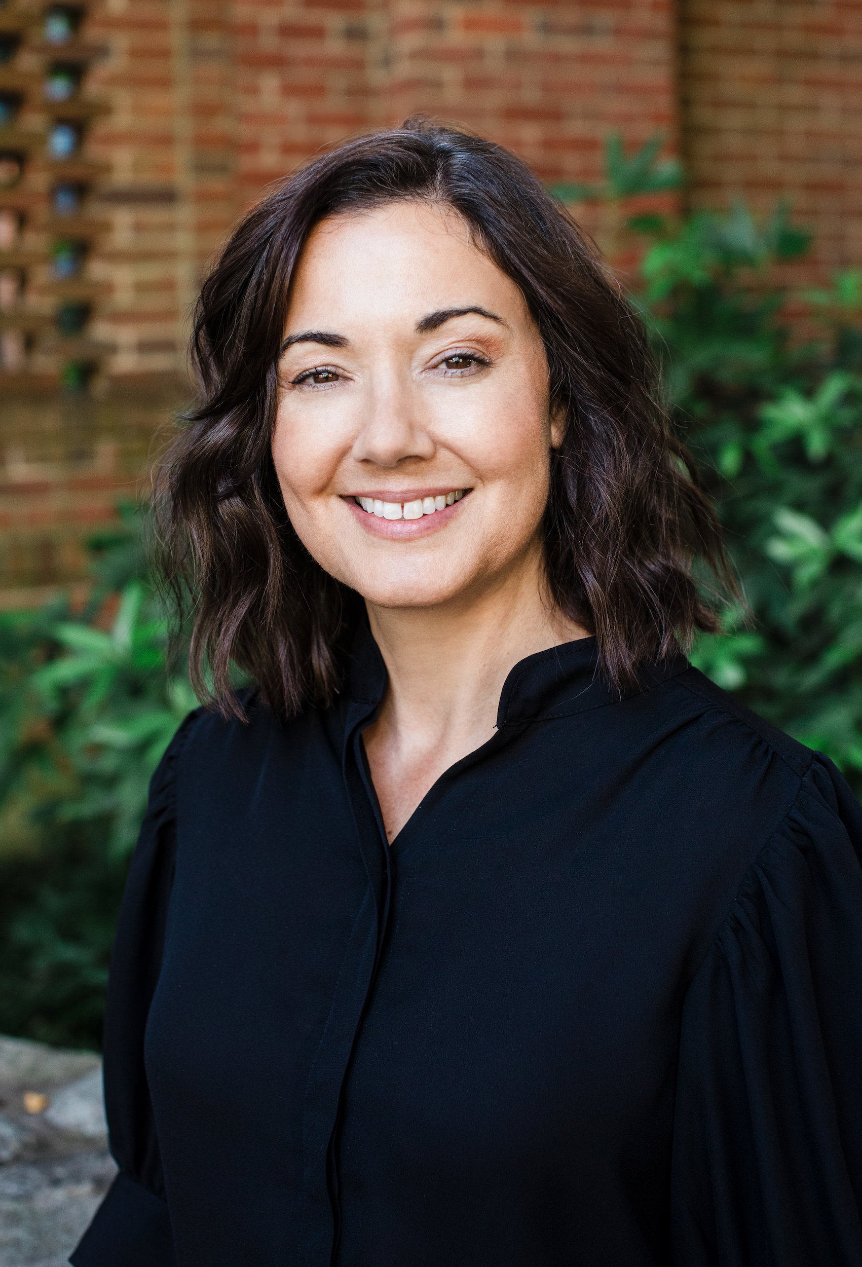 Kristen Calhoun smiles in a professional outdoor portrait against a textured brick wall.
