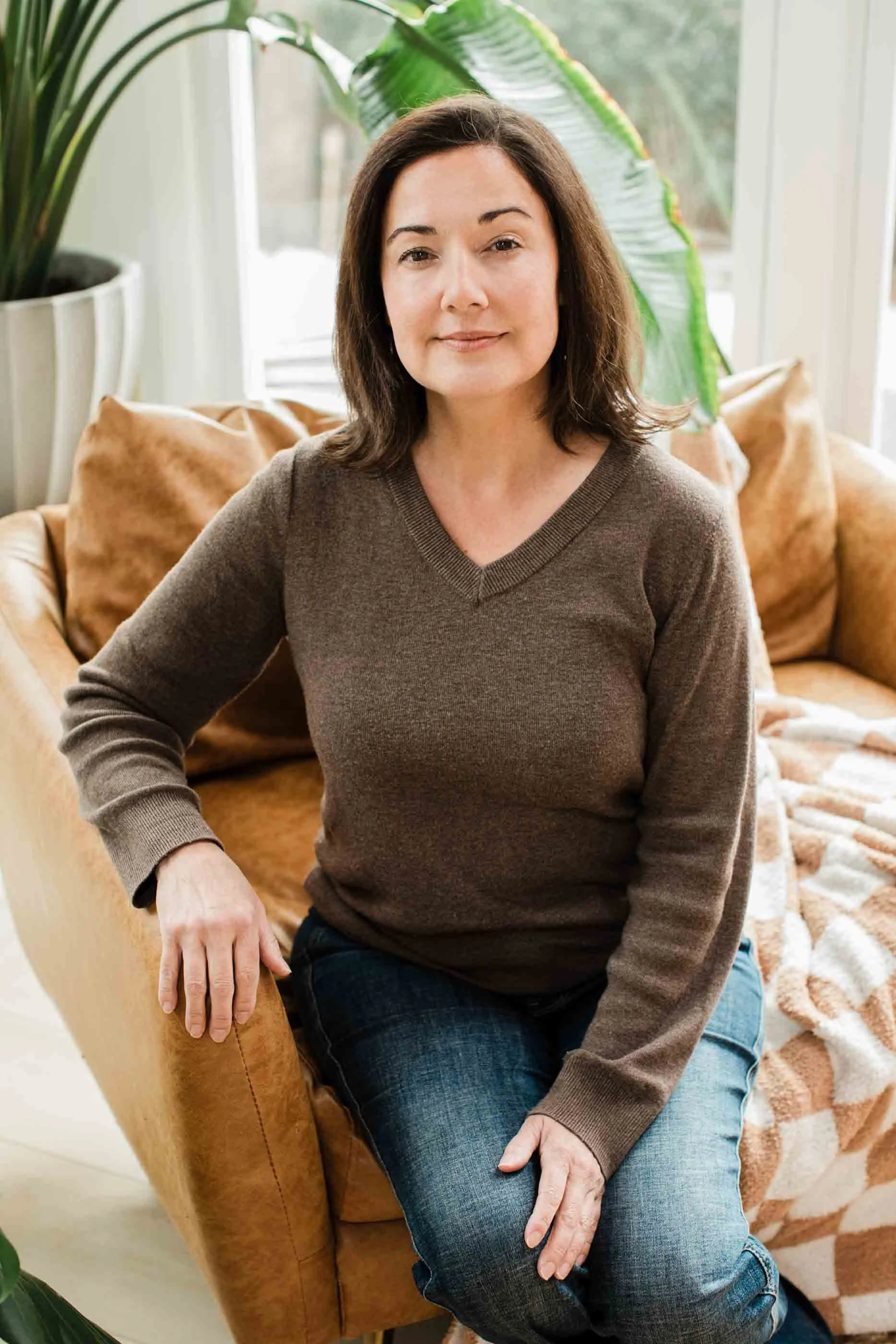 Woman in brown V-neck sweater and blue jeans sitting on tan leather couch near window and houseplants.
