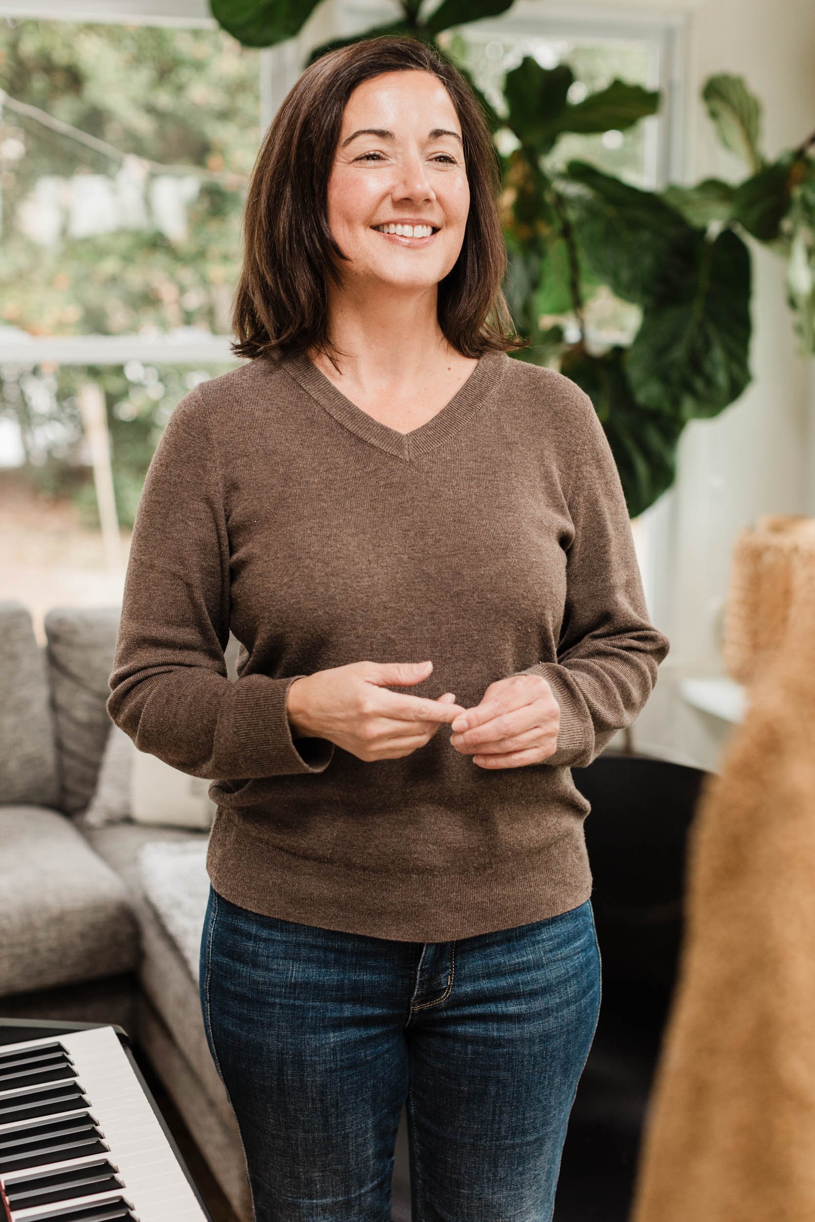A woman smiling, standing indoors near a window with greenery outside, in a brown sweater and jeans, with a piano partially visible on the left.
