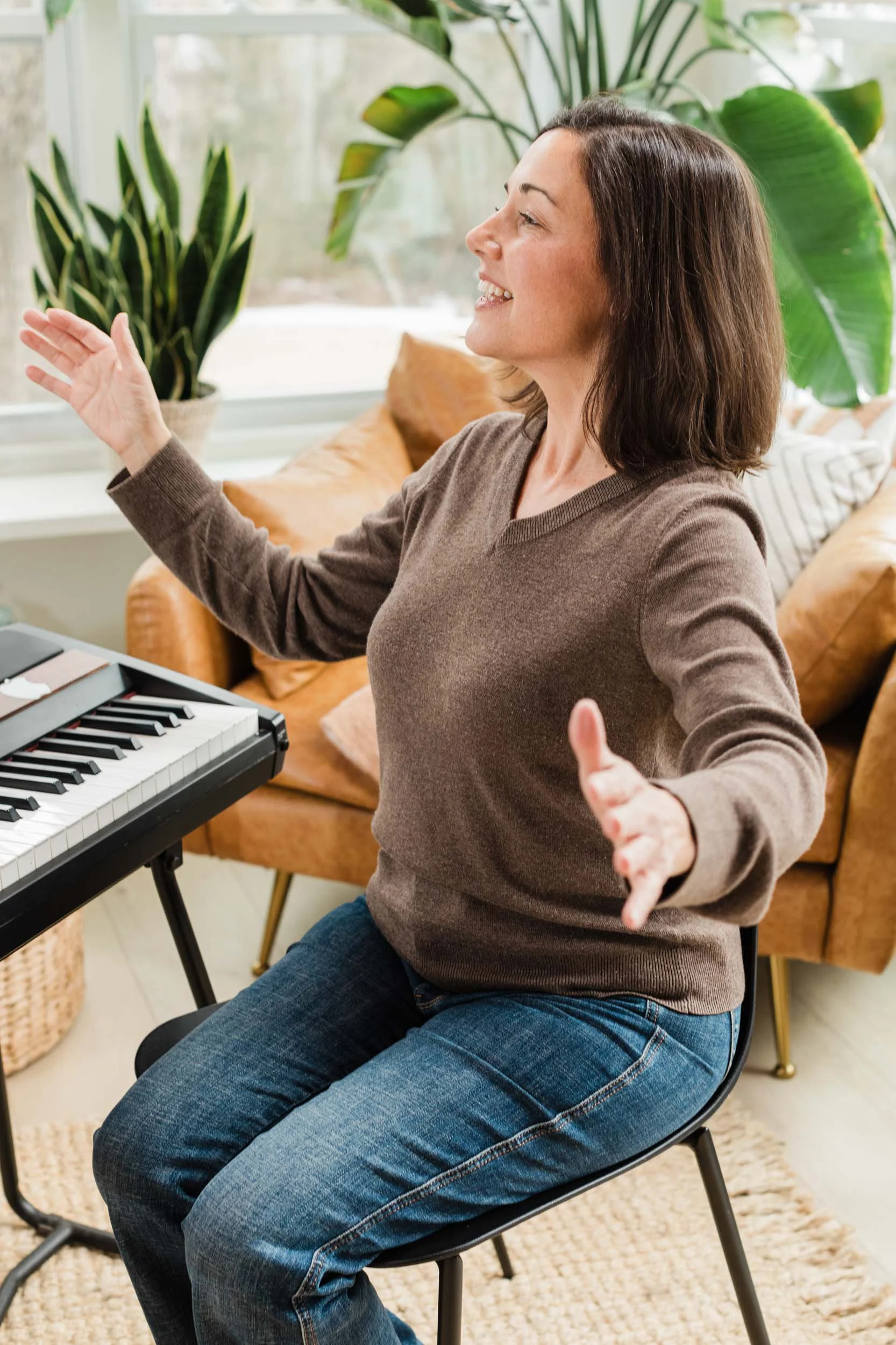Kristen Calhoun smiling and gesturing with open arms while sitting at a keyboard in a sunny living room.