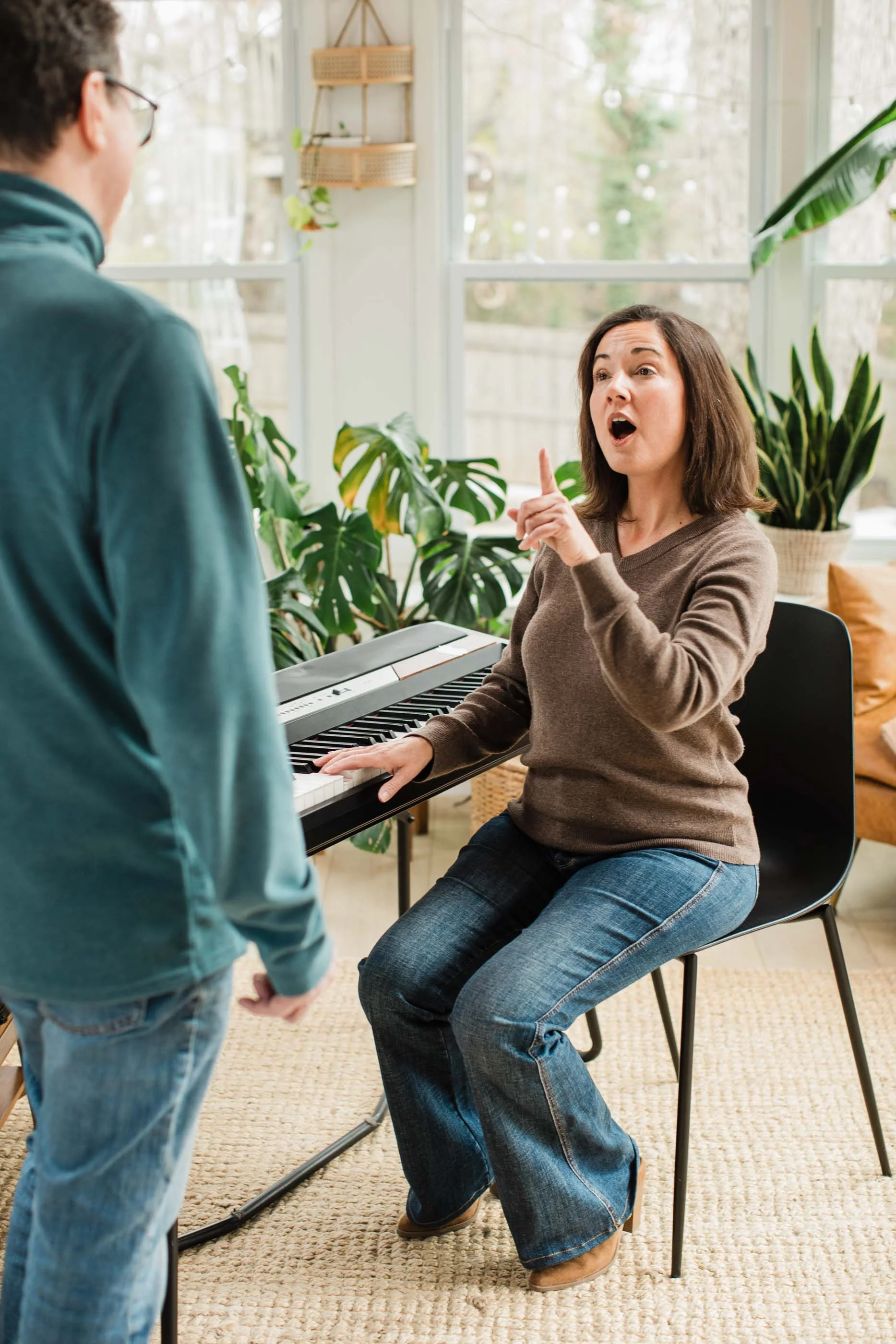 Kristen Calhoun sings at a keyboard, gesturing to a listening student in a room with large windows, plants, and natural light.