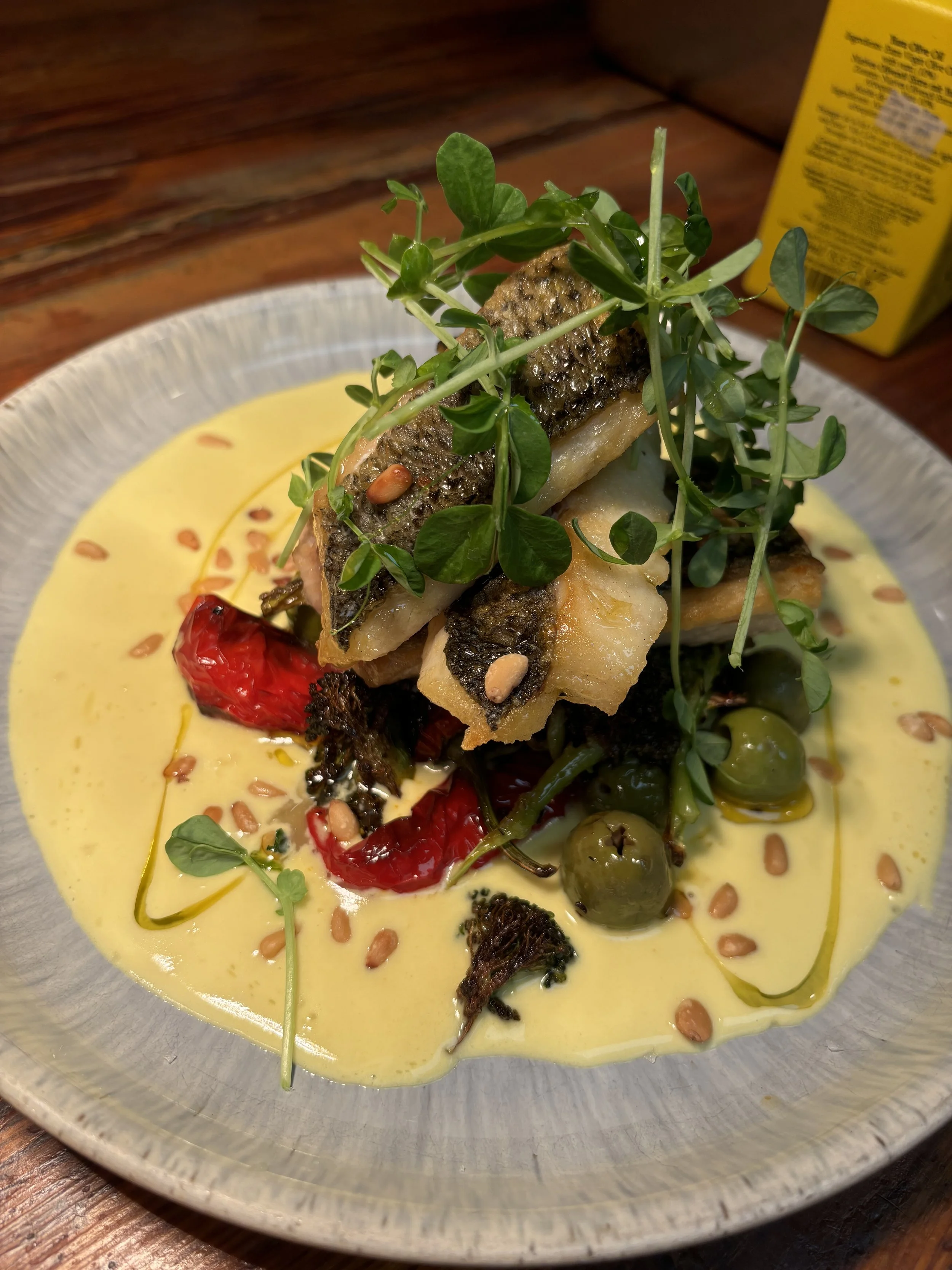 Dish with grilled fish placed on a bed of green herbs and peppers, drizzled with a yellow sauce and garnished with pine nuts and microgreens.
