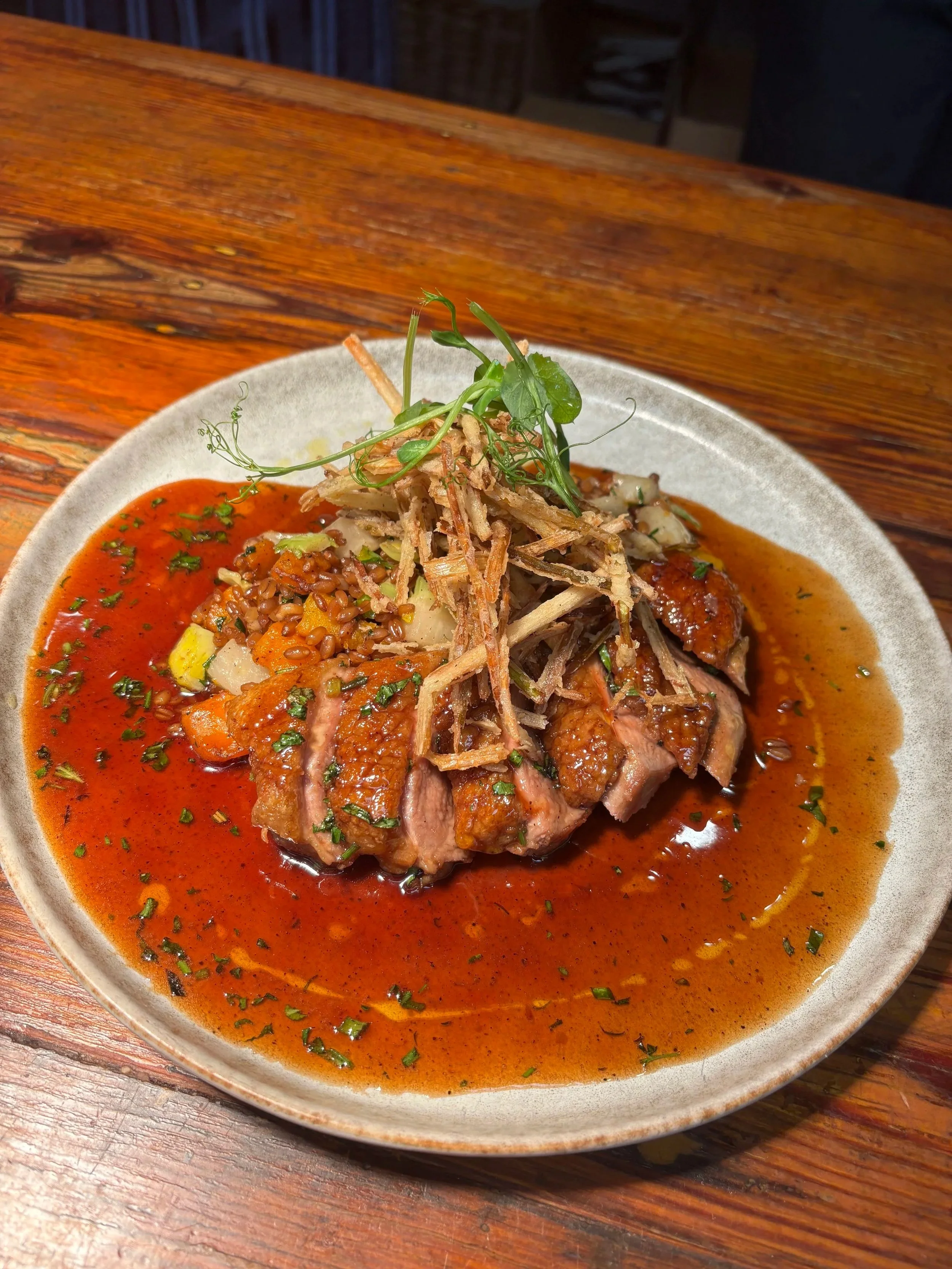 A plated dish featuring sliced cooked meat topped with crispy fried onions and garnished with microgreens, served with a rich brown gravy and rice with vegetables on a rustic wooden table.