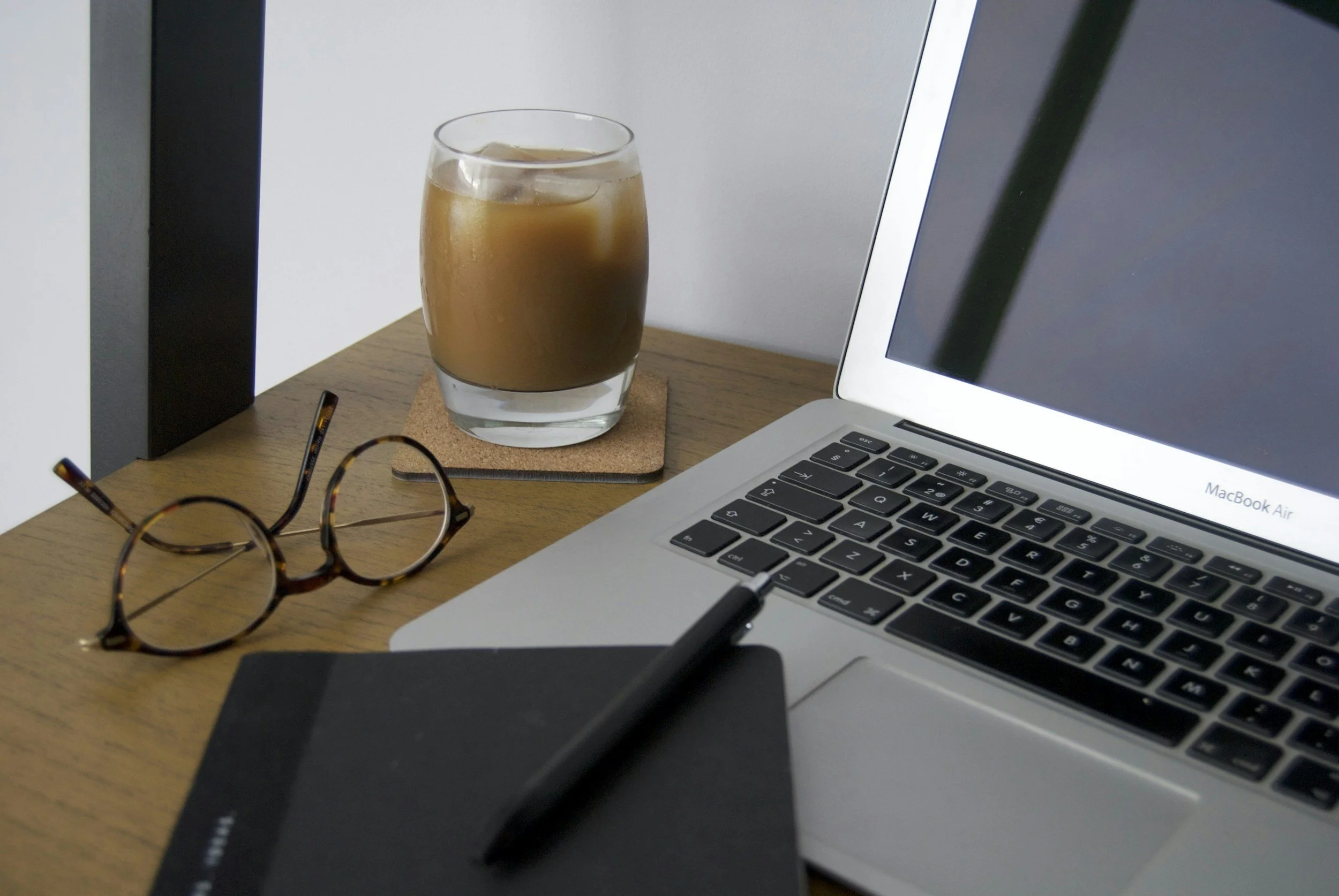 A wooden desk with a MacBook Air laptop, a black drawing tablet with a stylus, a pair of tortoiseshell glasses, a glass of iced coffee on a cork coaster, and a closed notebook.