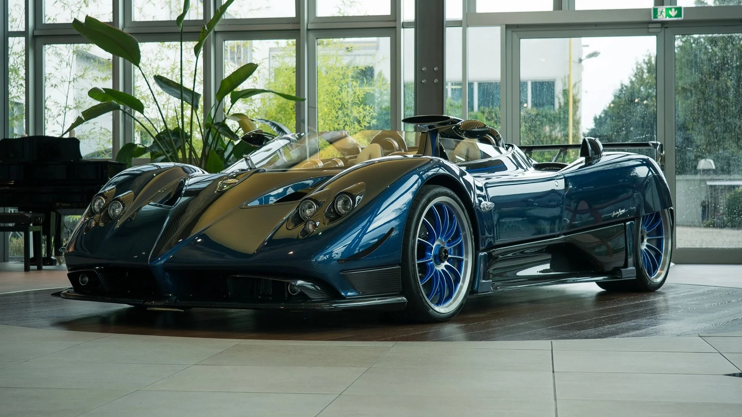A blue sports car displayed indoors near large windows with plants and a piano in the background.