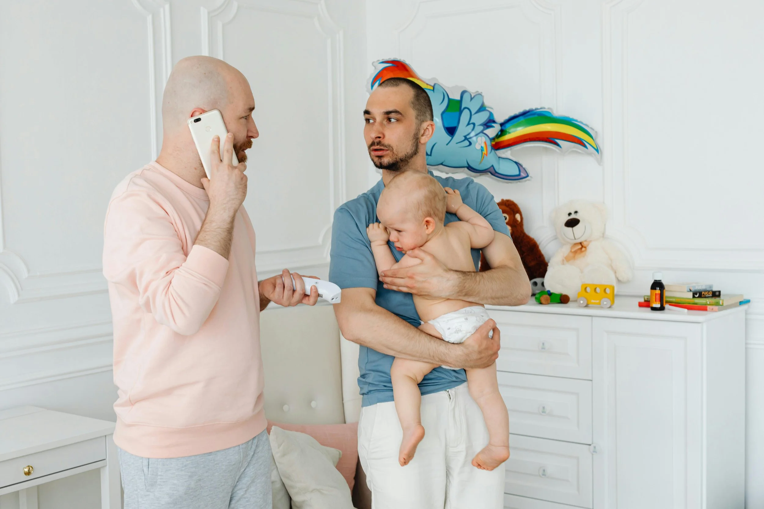 Two fathers in a nursery caring for their baby, highlighting inclusive parenting and family mental health support.