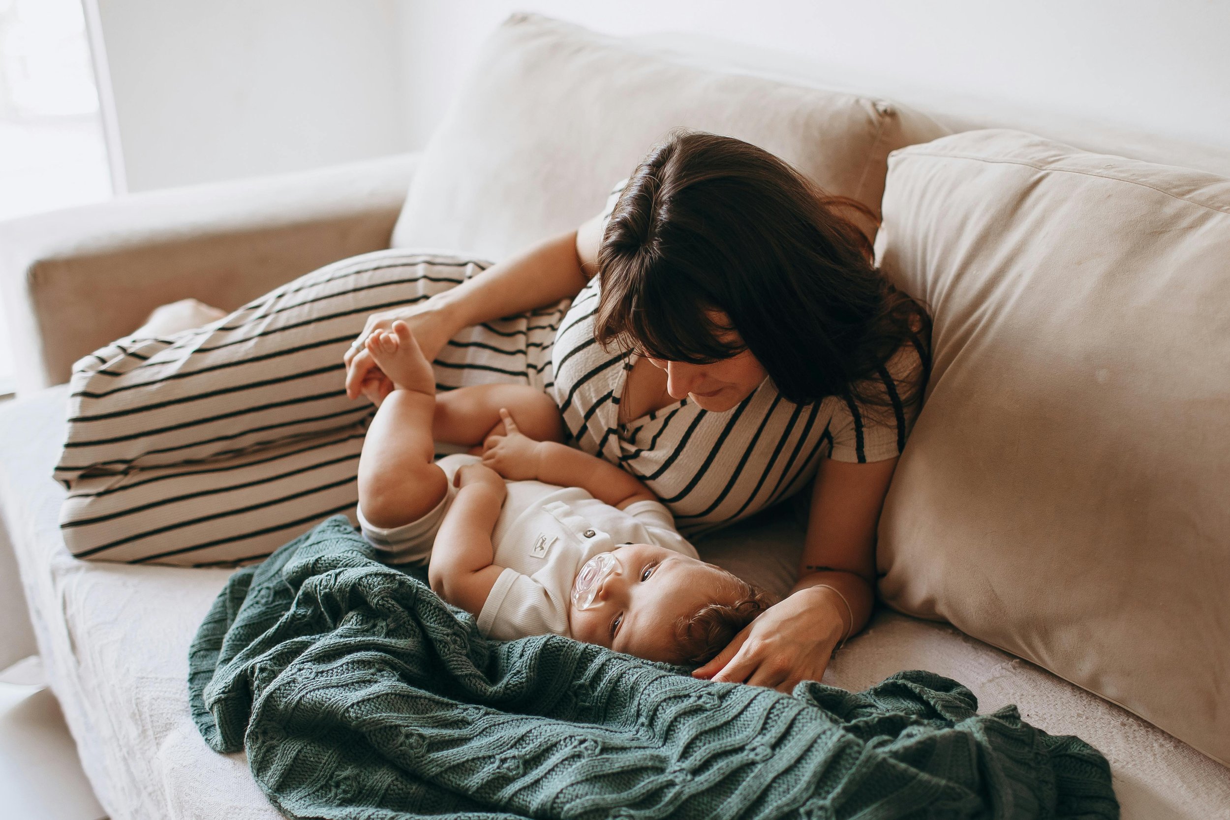 Mother resting with baby on sofa in warm sunlight — early postpartum connection and healing.