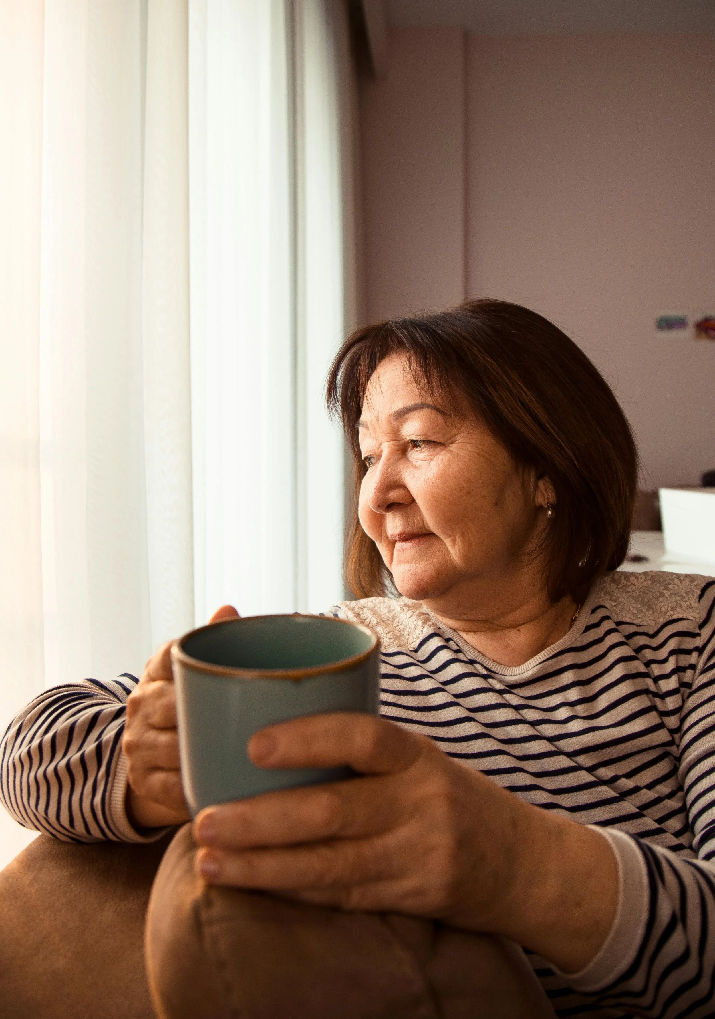 Older woman sitting by window with mug — life reflection, identity transitions, and emotional wellbeing.