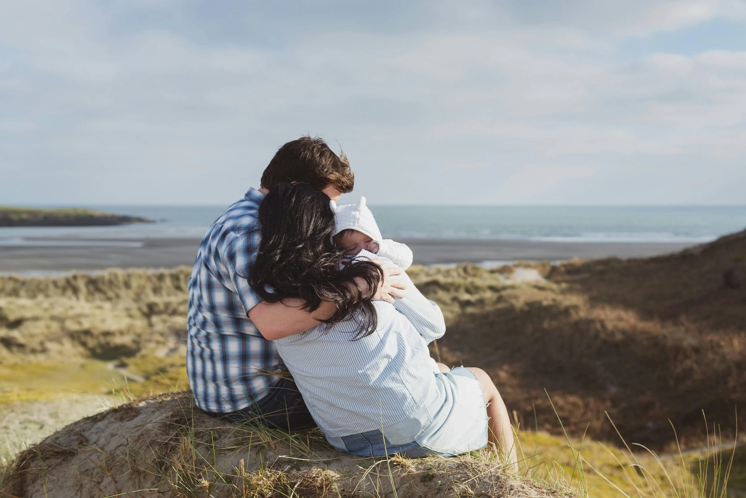 Family with baby sitting on coastal hill — family bonding and life transition after birth.